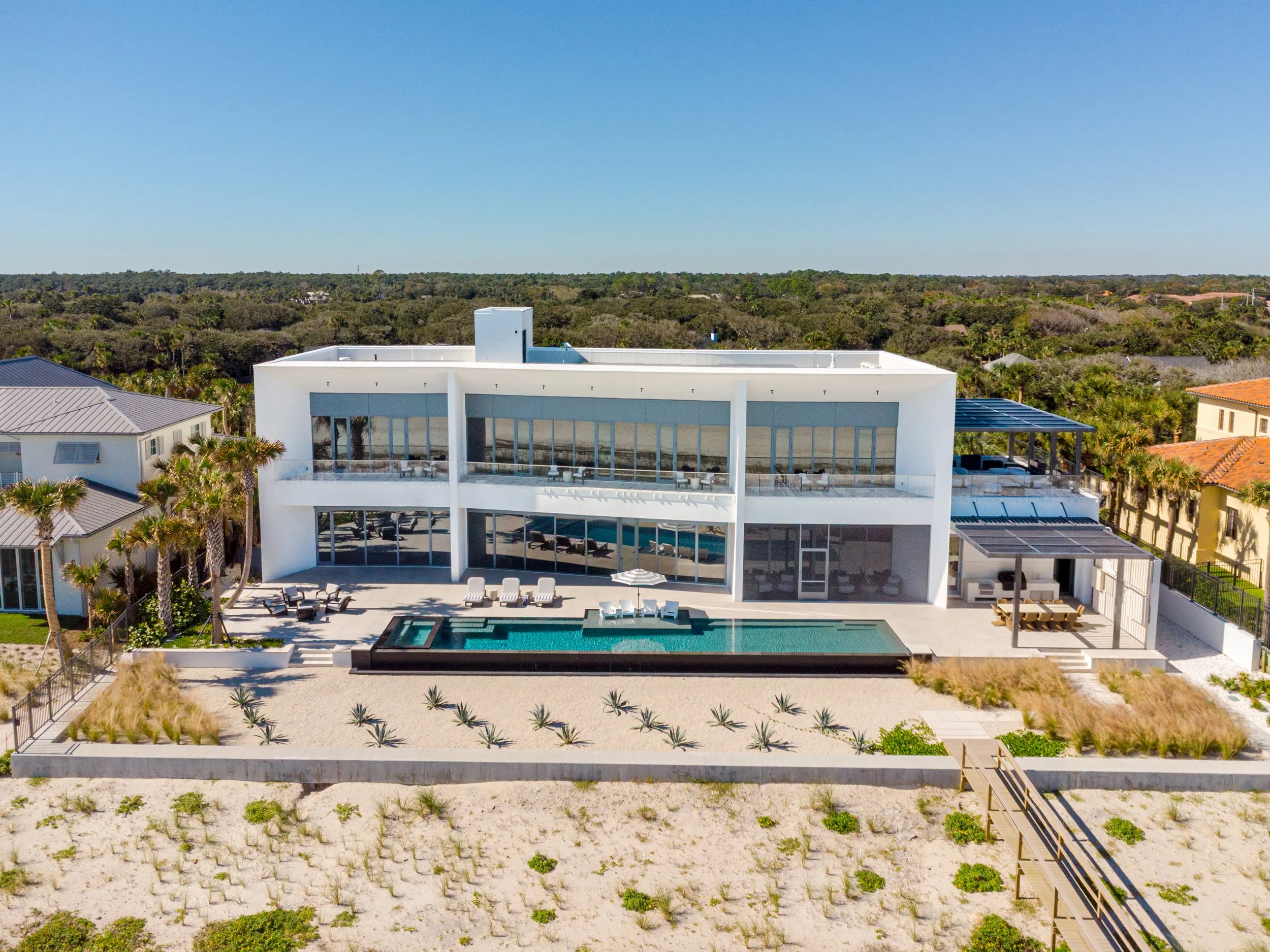 Aerial view of modern white house with large glass windows, rooftop pool, patio, and surrounding desert landscape with palm trees and neighboring houses.