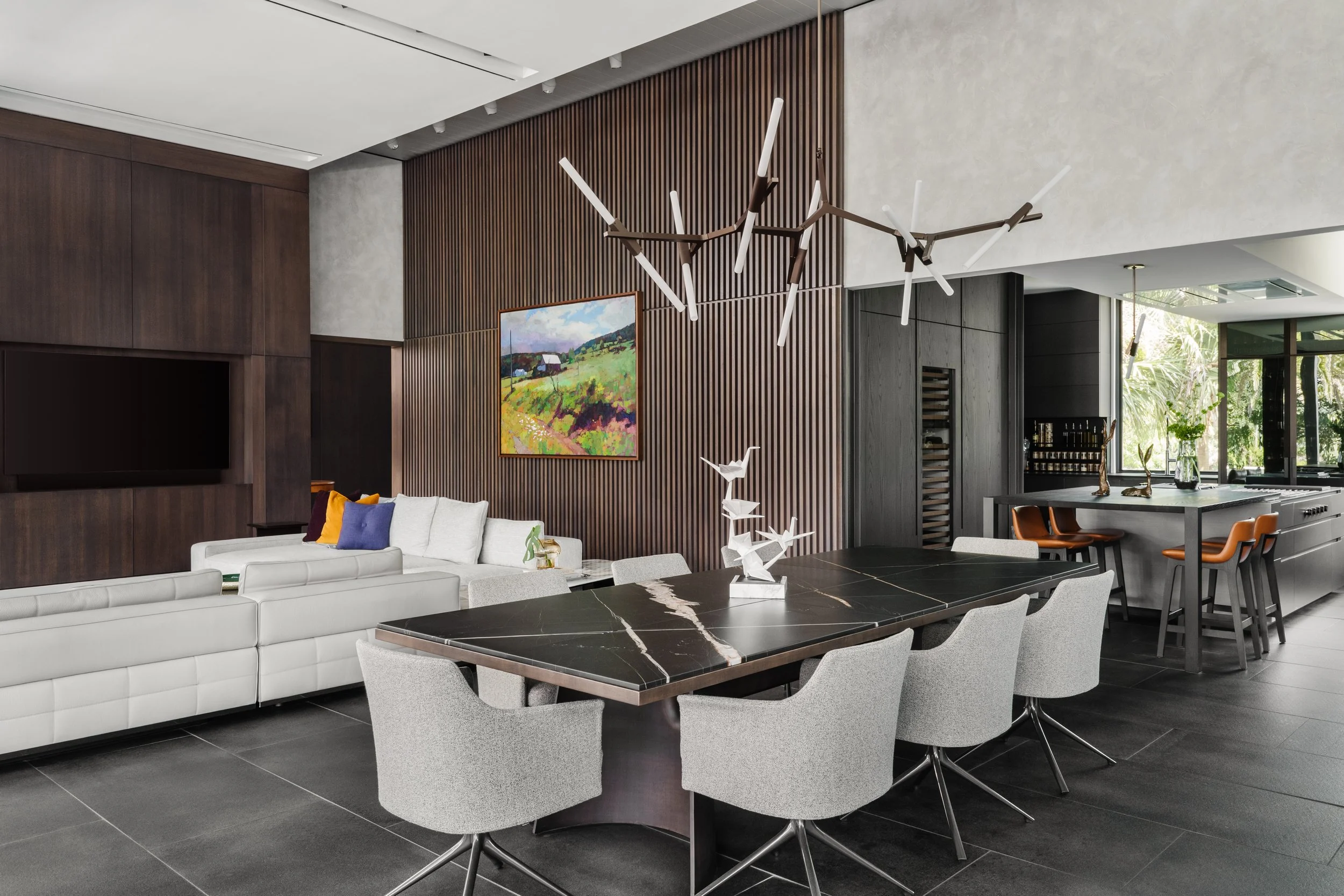 Modern living and dining area featuring a white sectional sofa with colorful pillows, a black marble table, light gray chairs, a dark wooden wall with vertical slats, colorful landscape painting, contemporary chandelier, and large windows with a view