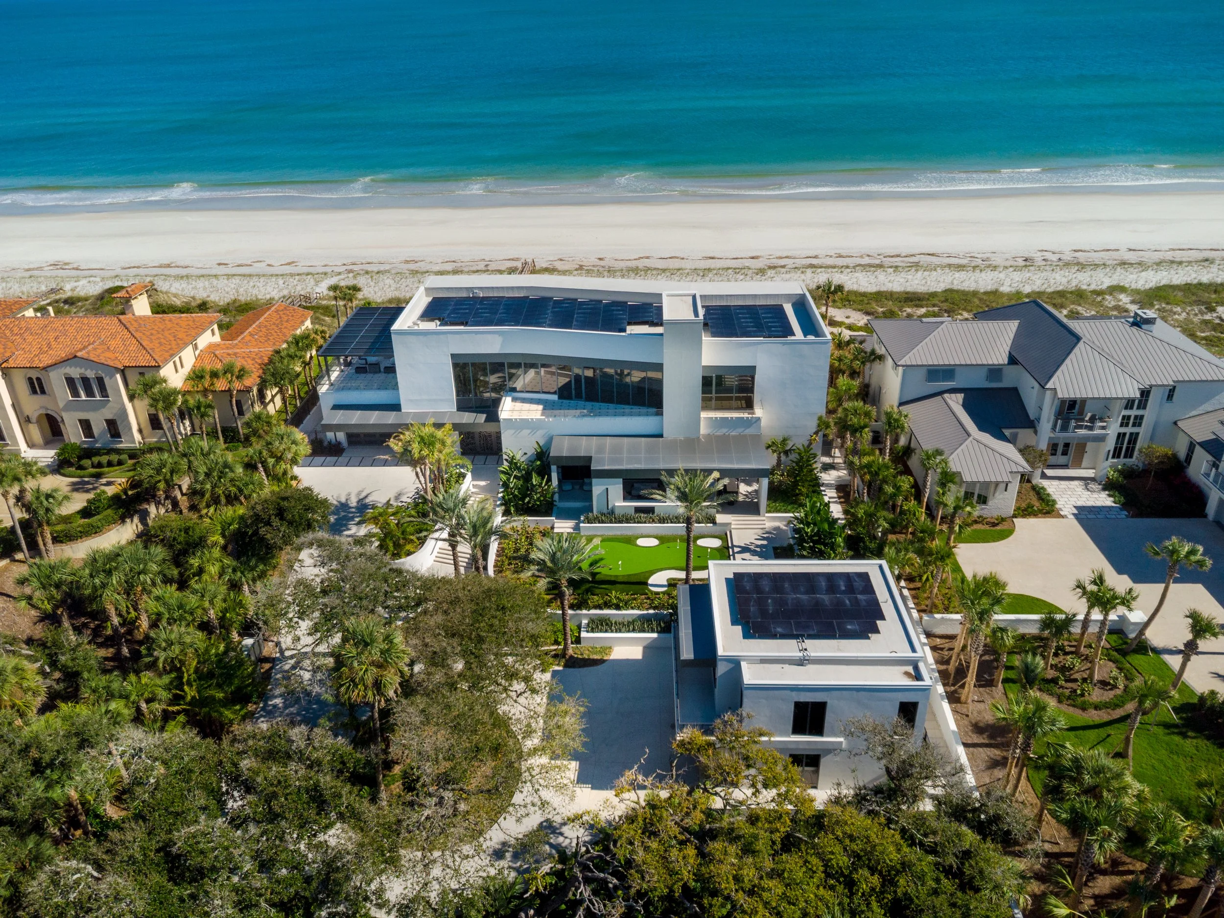Aerial view of beachfront houses with solar panels on roofs, lined along a sandy beach and blue ocean.