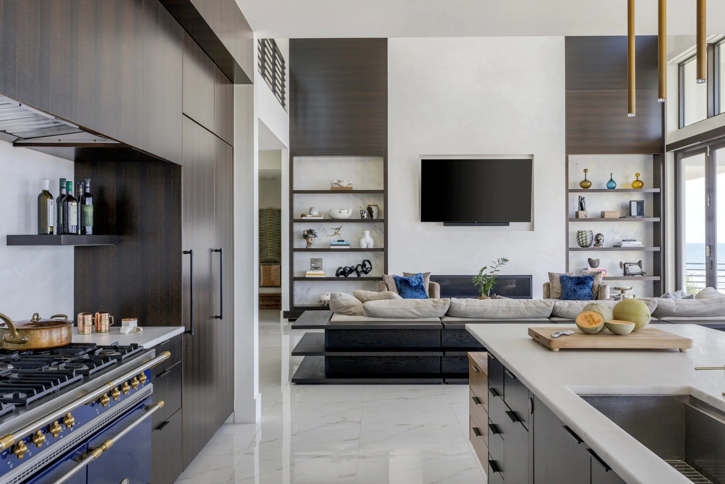 Open-concept modern living space with a kitchen, shelves with decorative items, a wall-mounted TV, and a comfortable seating area with cushions and plants.