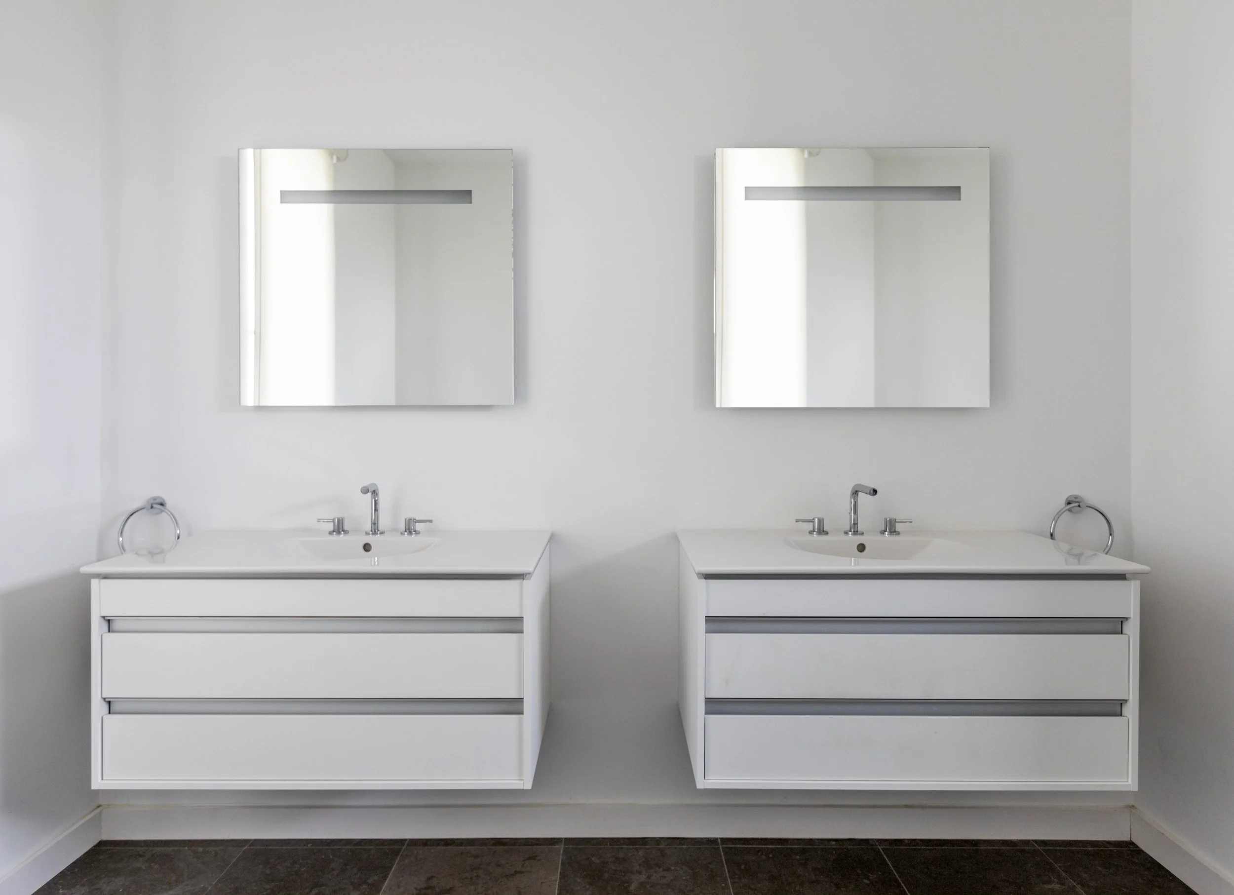 Double bathroom vanity with two white sinks, two faucets, and two mirrors on white wall, with dark tiled floor.