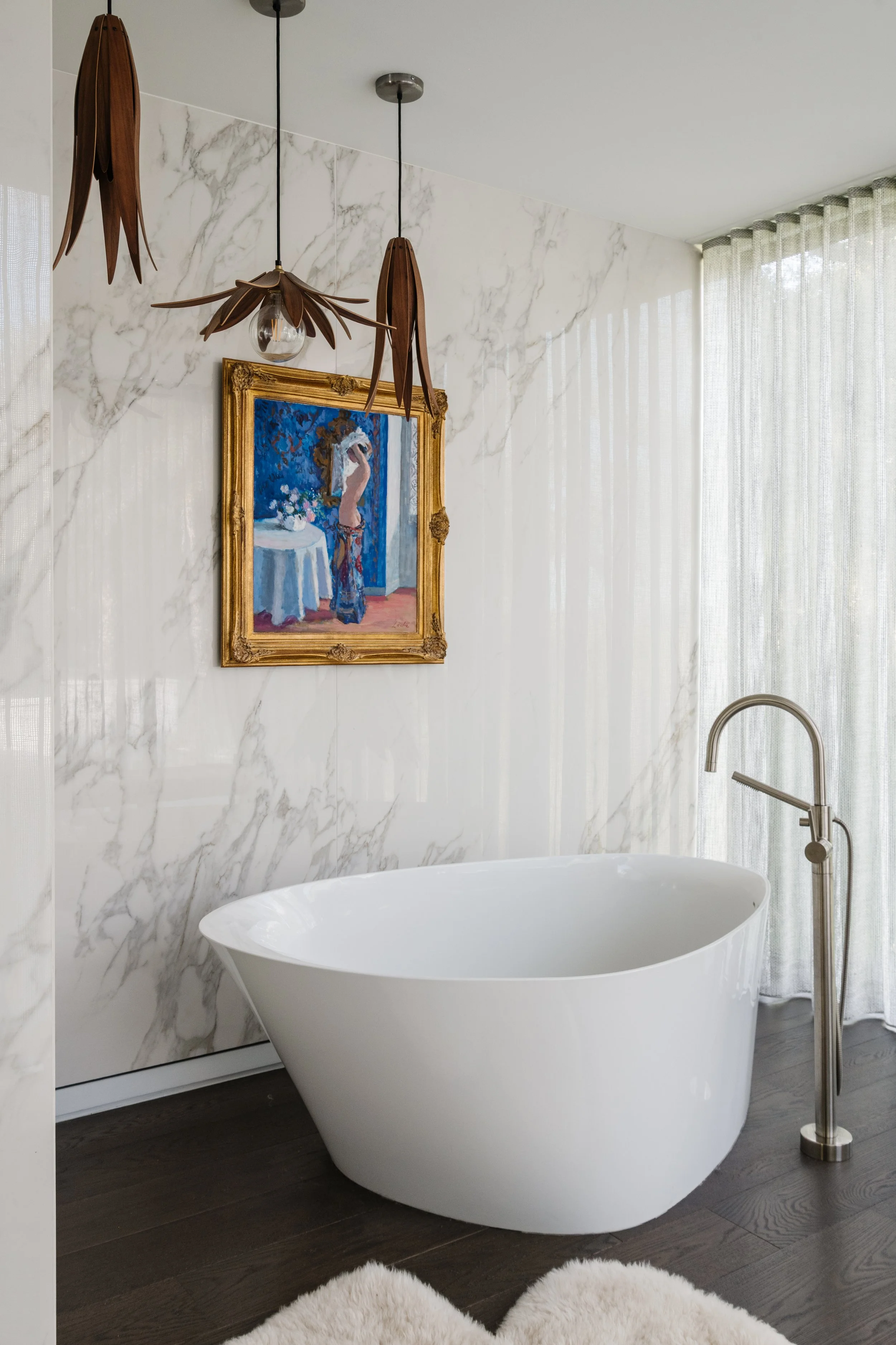 A modern bathroom with a white freestanding bathtub, dark wood flooring, a marble wall, sheer curtains, a gold-framed painting of a woman, and wooden pendant lights.