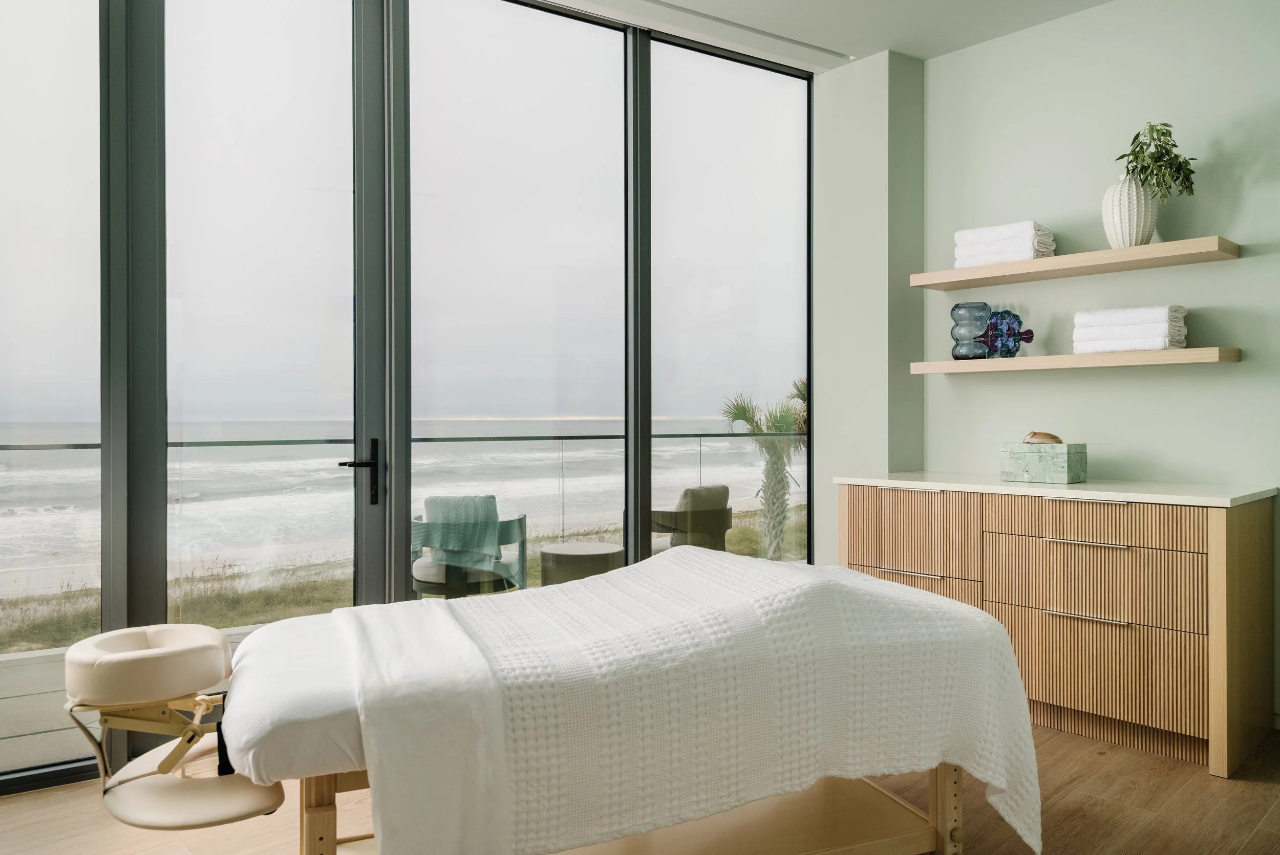 A spa or massage room with a massage table covered in a white blanket, a beige massage chair nearby, and large glass sliding doors revealing a beach and ocean view outside. There are two wooden shelves on the green wall holding folded white towels, a