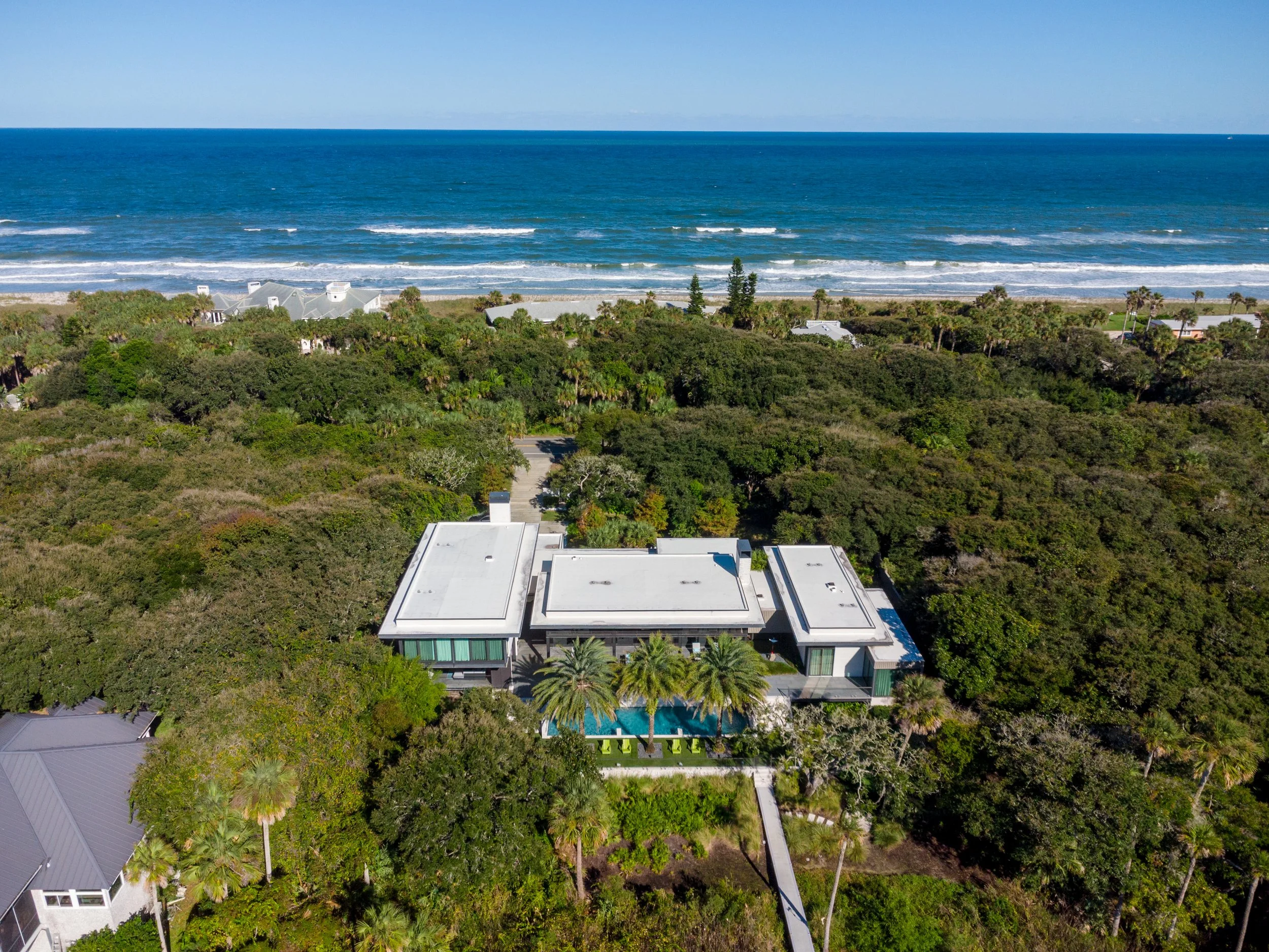 Aerial view of a coastal area with houses and lush green trees near the beach and ocean, under a clear blue sky.