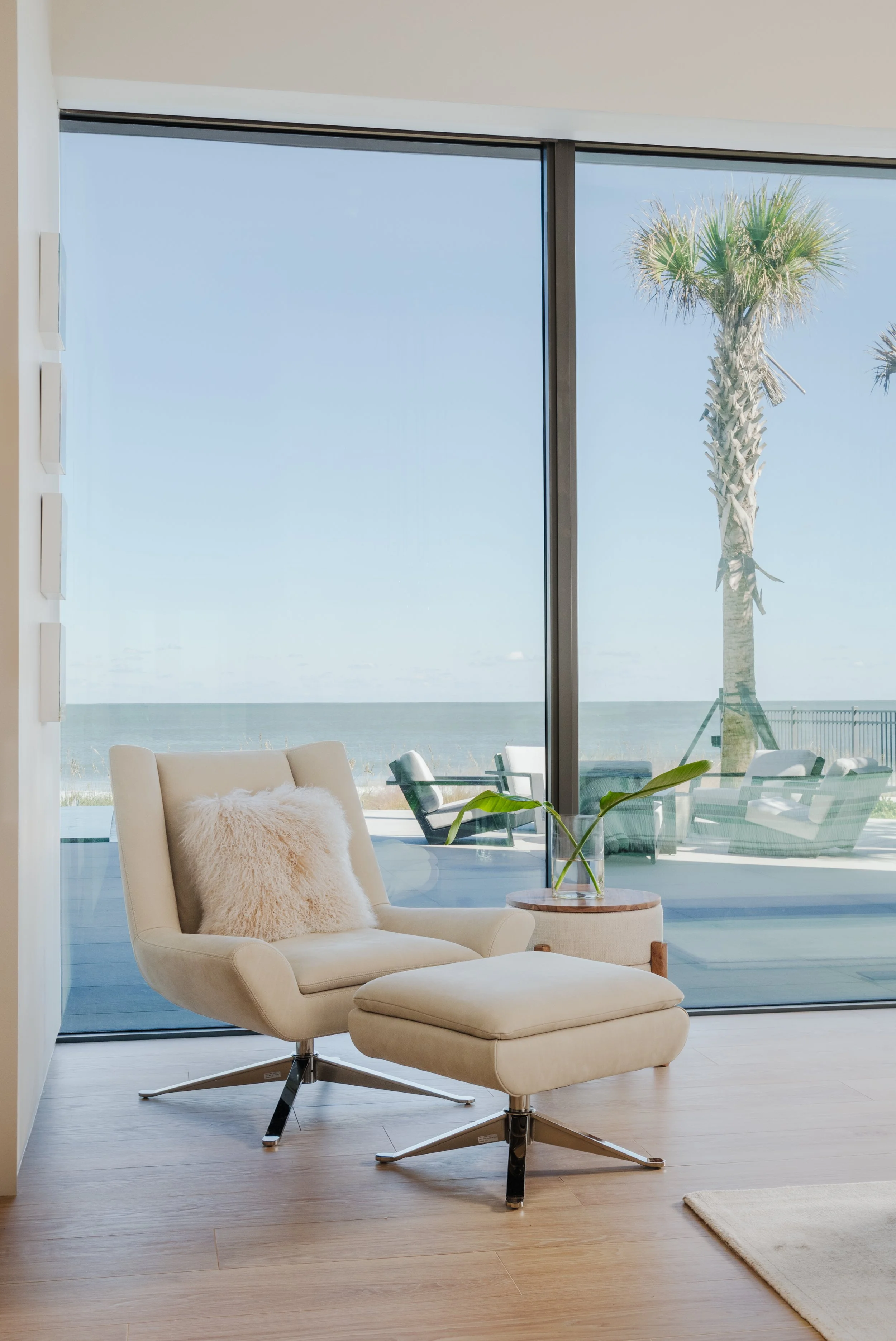 Modern living space with a cream swivel lounge chair with matching ottoman, a fluffy cream pillow, a glass vase with a green plant, large floor-to-ceiling window showing palm trees and the ocean, and wood flooring.