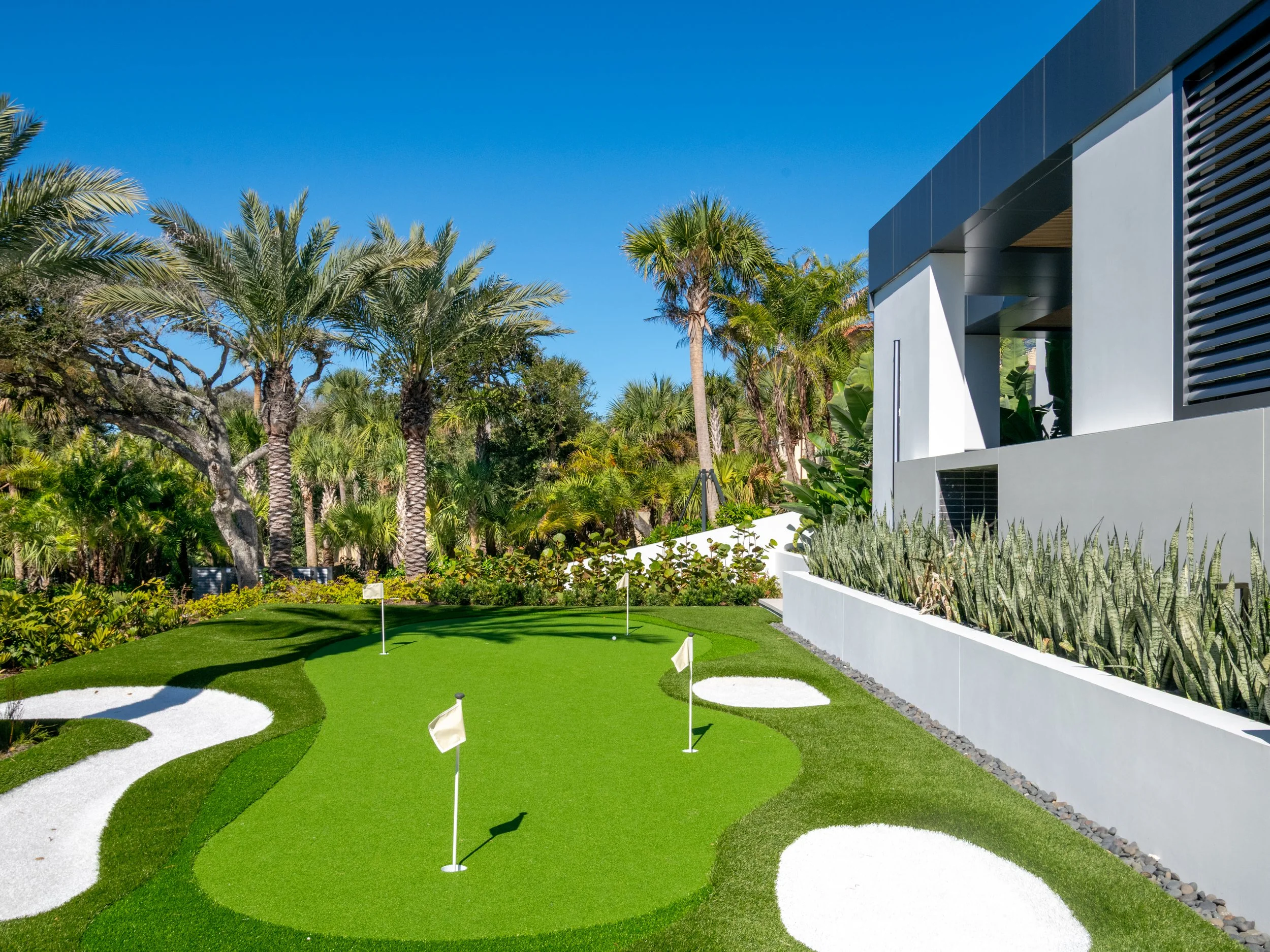 Modern house with white exterior surrounded by tall palm trees and lush greenery, featuring a mini golf course with flags and sand traps in the yard, under a clear blue sky.