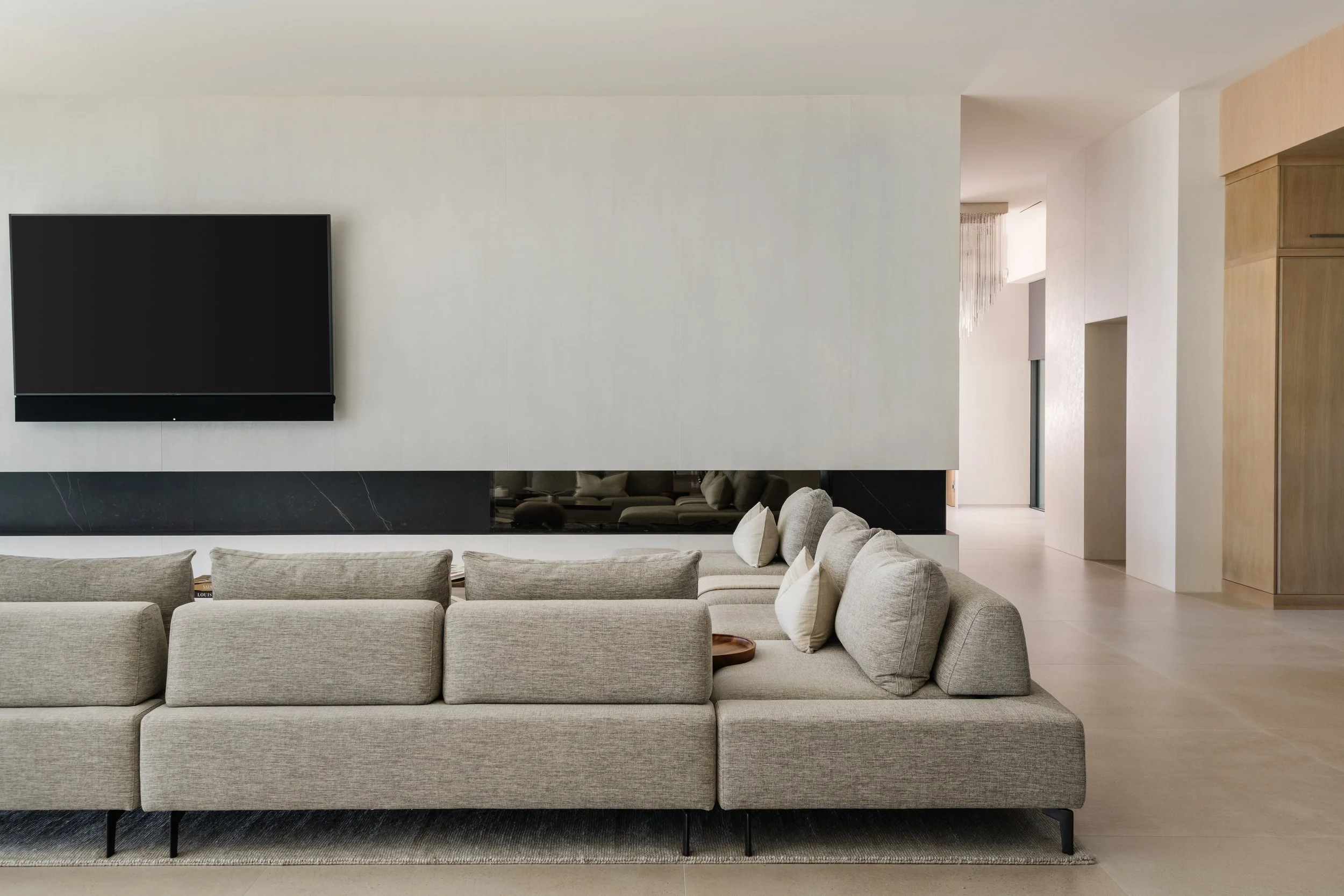 Modern living room with a large beige sectional sofa, mounted flat-screen TV on a white wall, and a wooden cabinet on the right side.