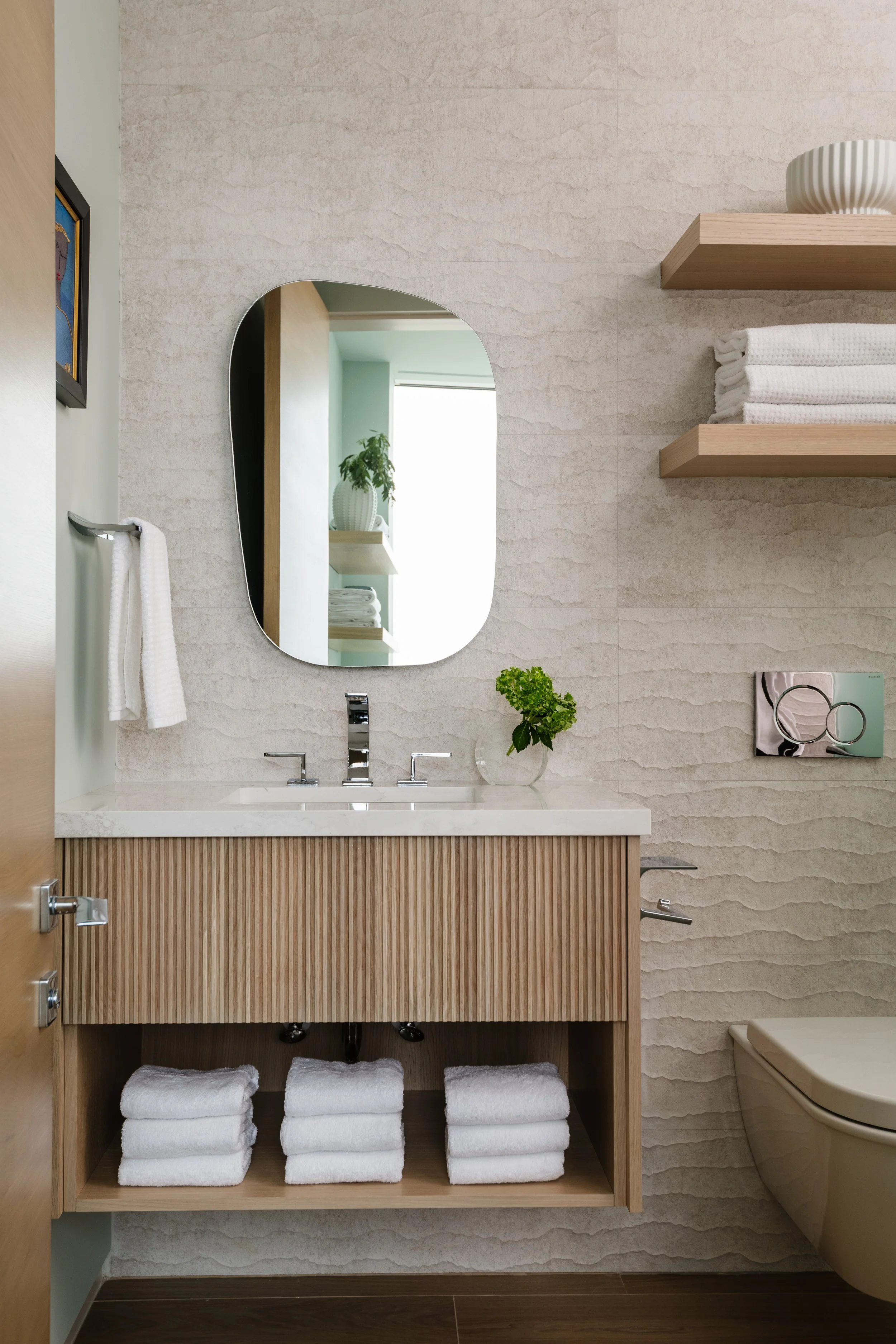 Bathroom with minimalist design featuring a wooden vanity with vertical slats, white countertop, and stacked white towels underneath. The wall has textured beige tiles, a small potted green plant, a modern faucet, and a large irregularly shaped mirro