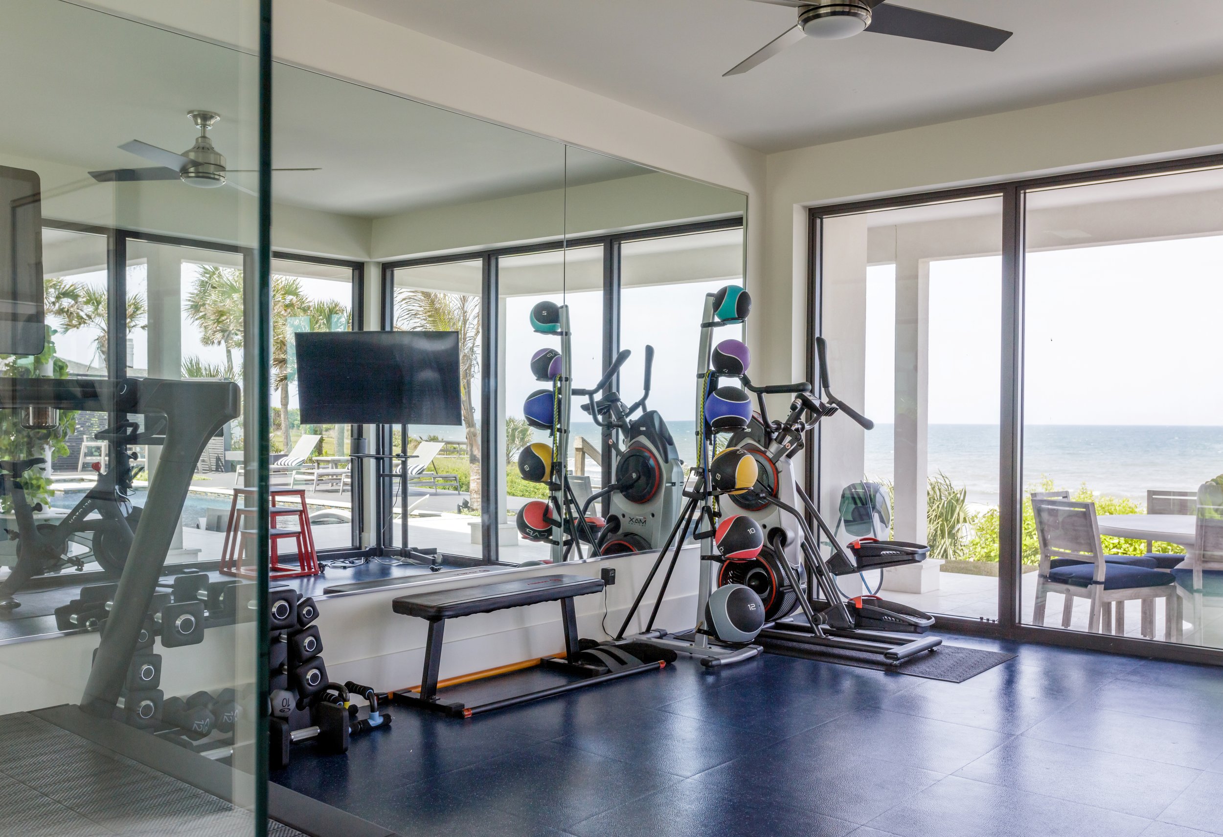 A modern gym with exercise equipment, large windows showing outdoor trees and ocean view, and a ceiling fan.