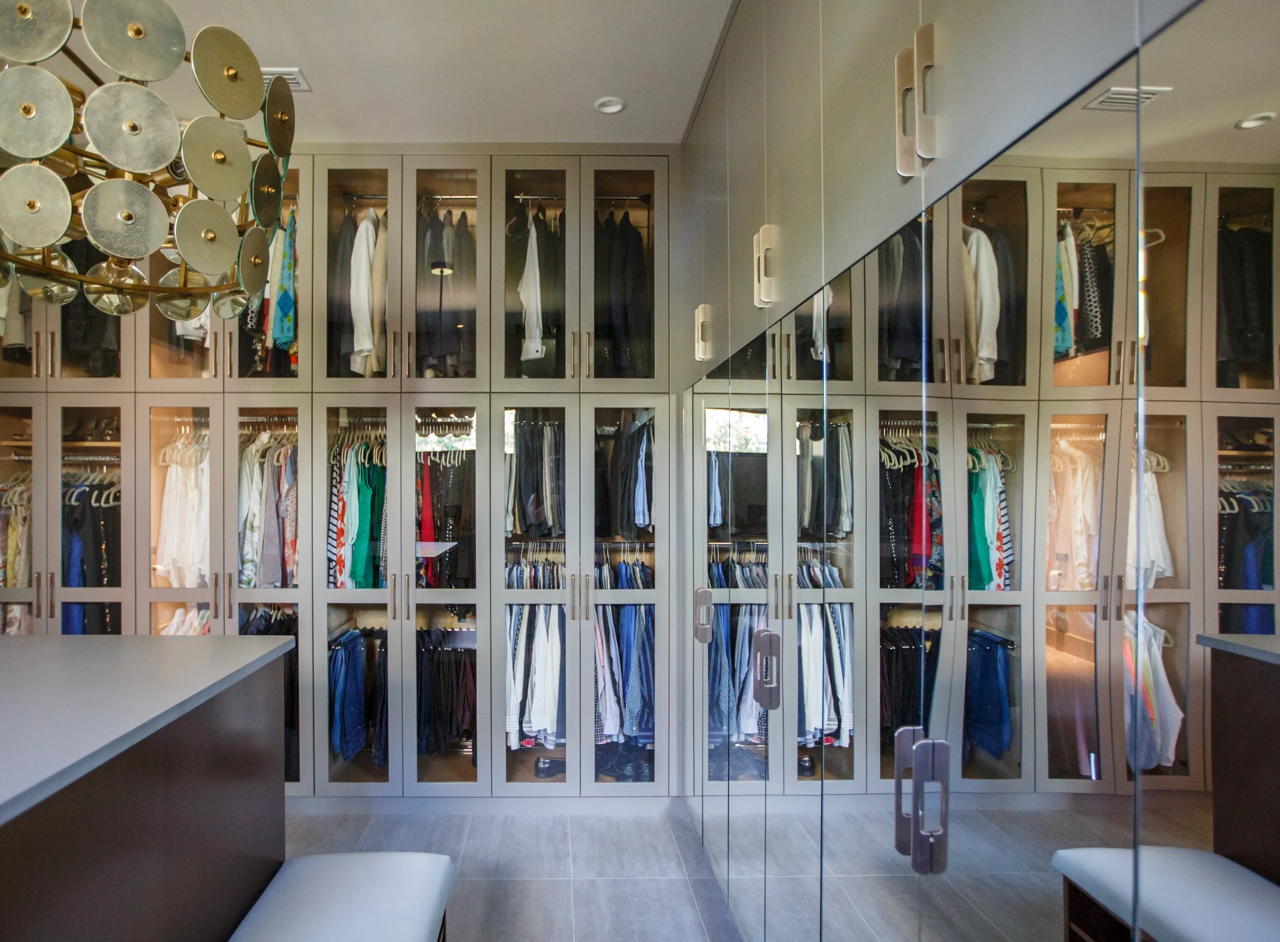 A walk-in closet with glass-front cabinets filled with clothes and shoes, a modern chandelier, and a mirrored sliding door. Seating with white cushions is visible in the foreground.