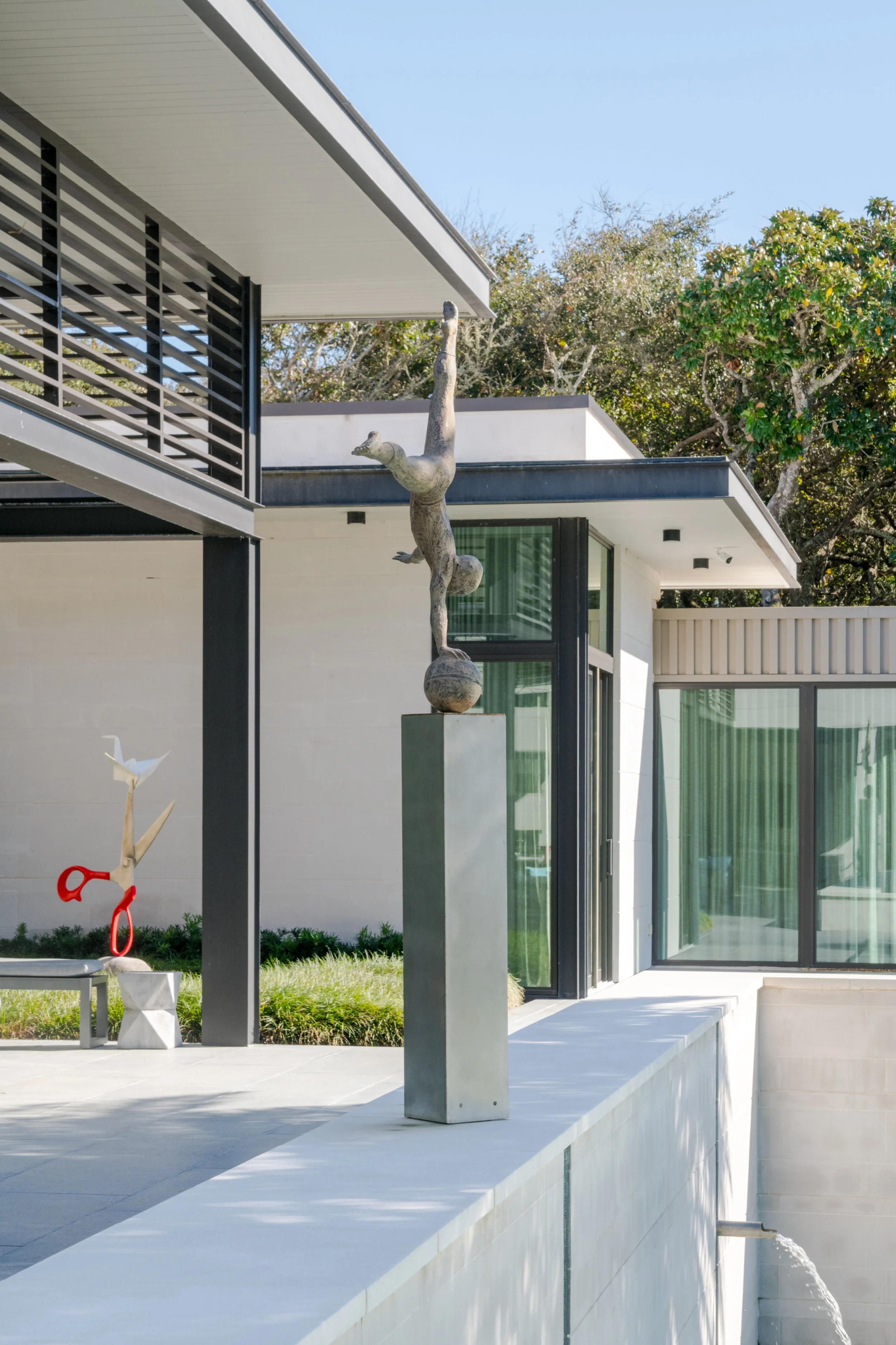 Modern house with large windows, an outdoor sculpture of a person balancing, and a small sculpture of white birds in the yard, under a clear blue sky.
