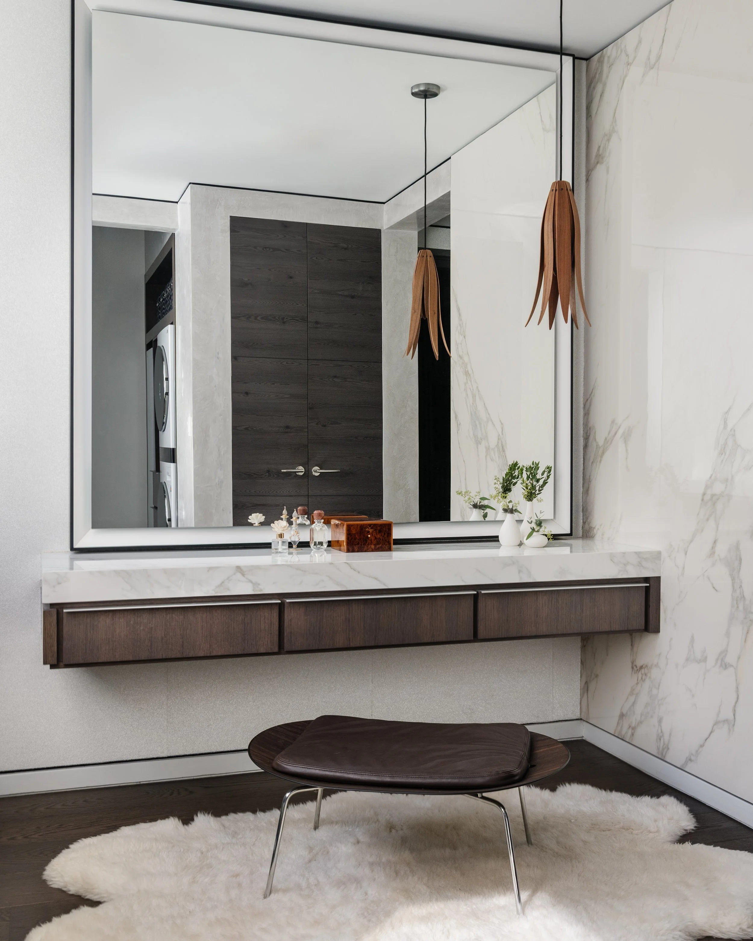 A modern minimalistic interior with a large wall mirror, a white marble countertop, a small potted plant, and decorative bottles. In front, a dark wooden bench with a round top and metal legs sits on a fluffy white rug, under a curtain. The wall behi