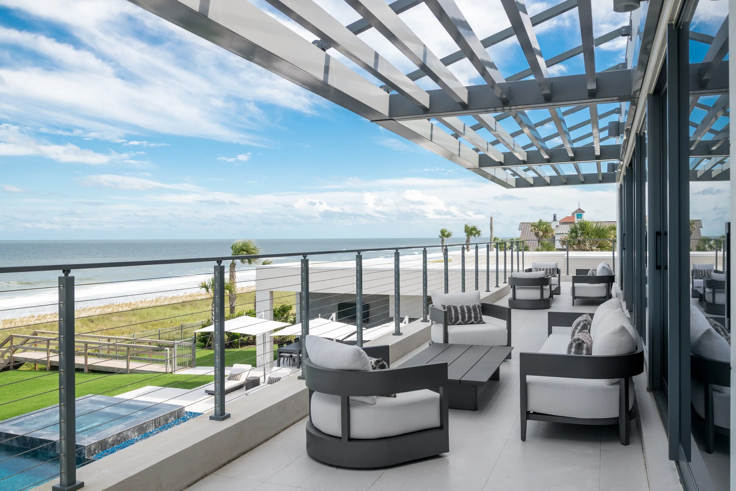 Balcony with white outdoor furniture, overlooking the beach, ocean, and cloudy sky, decorated with a metal pergola and glass railing.