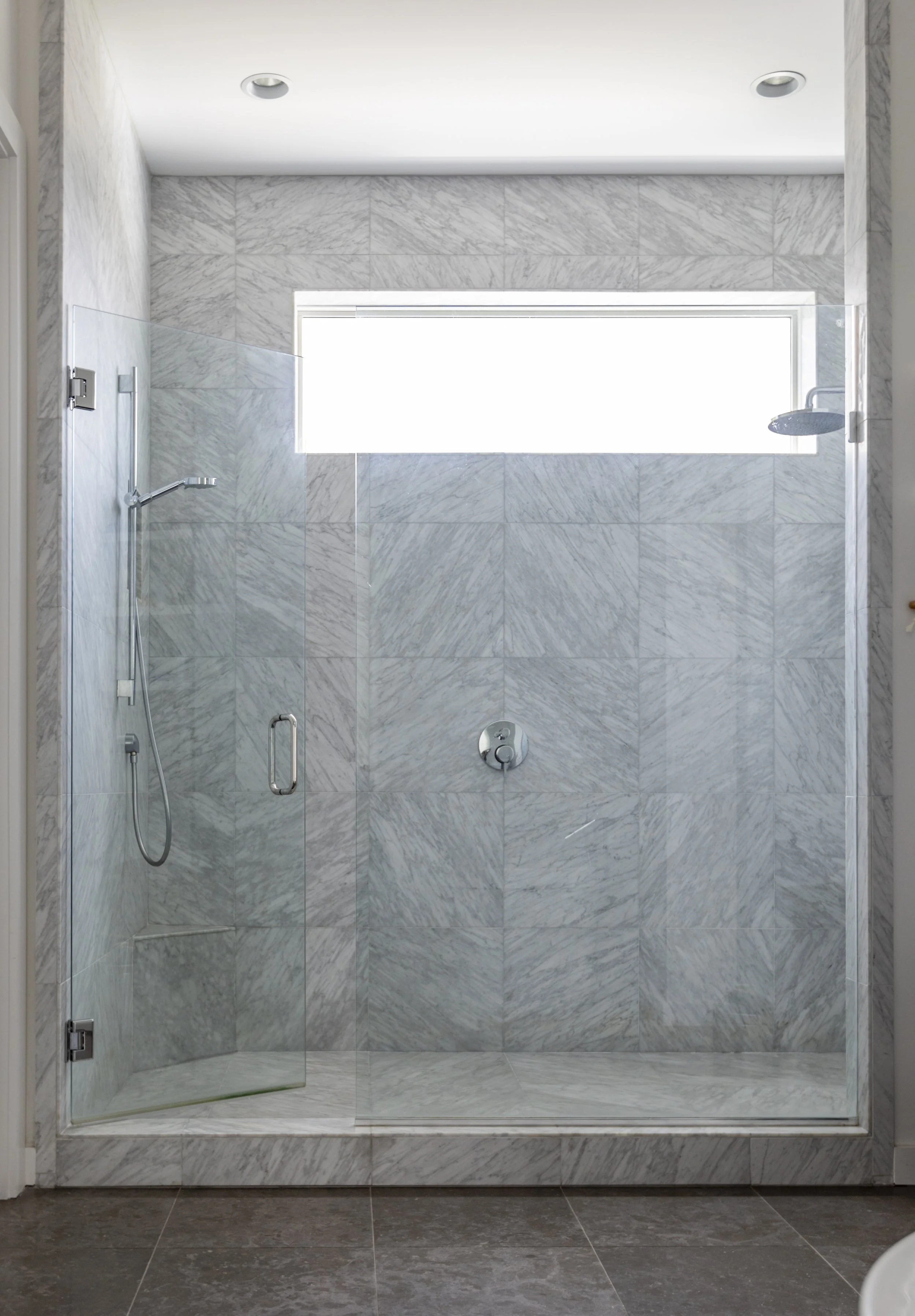 Modern walk-in shower with gray marble tiles, a large horizontal window, a glass door, a rain shower head, and a handheld shower wand.