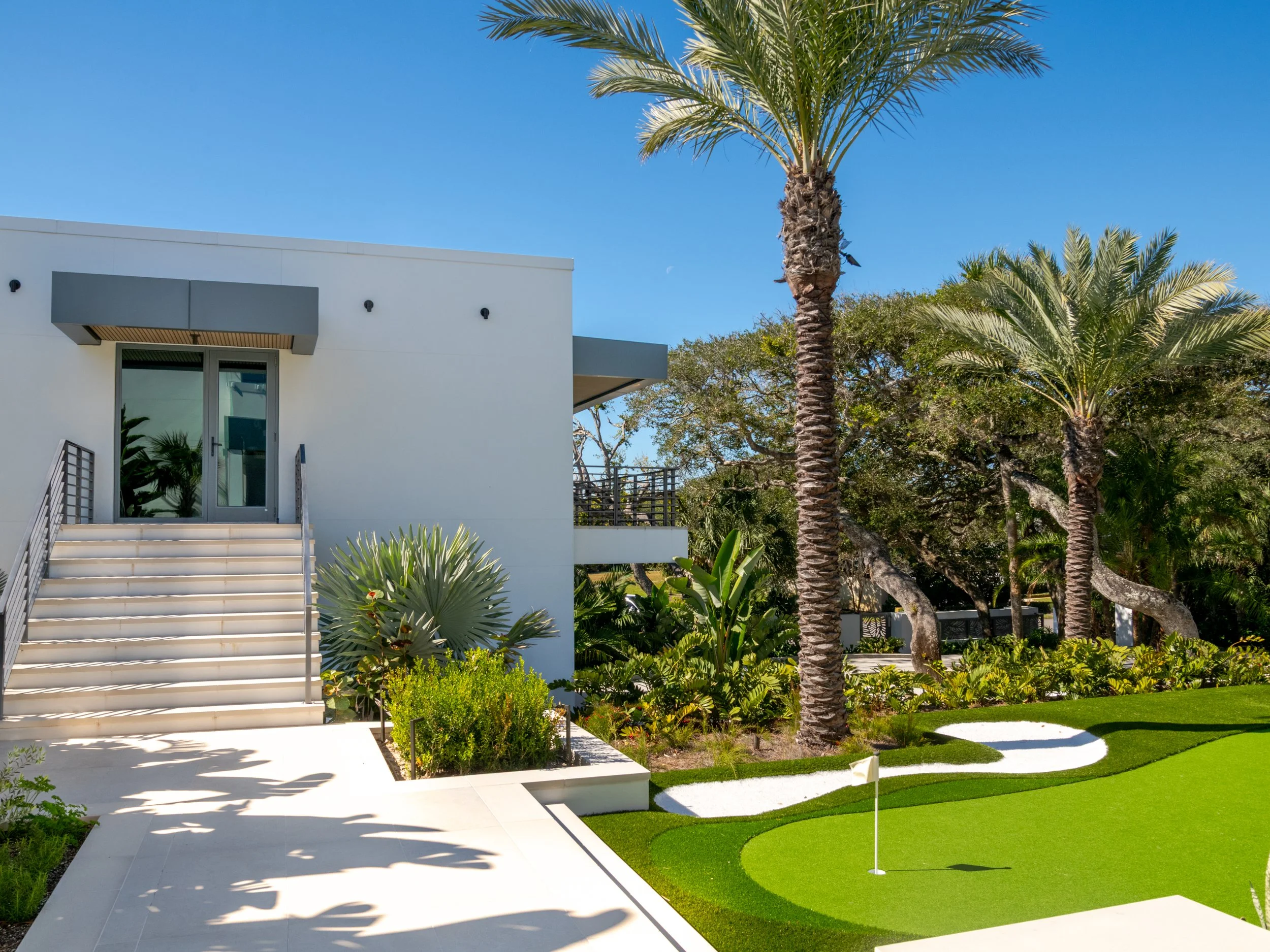 Modern white building with stairs leading up to a glass door, surrounded by a lush garden with palm trees, bushes, and a mini golf course under a clear blue sky.