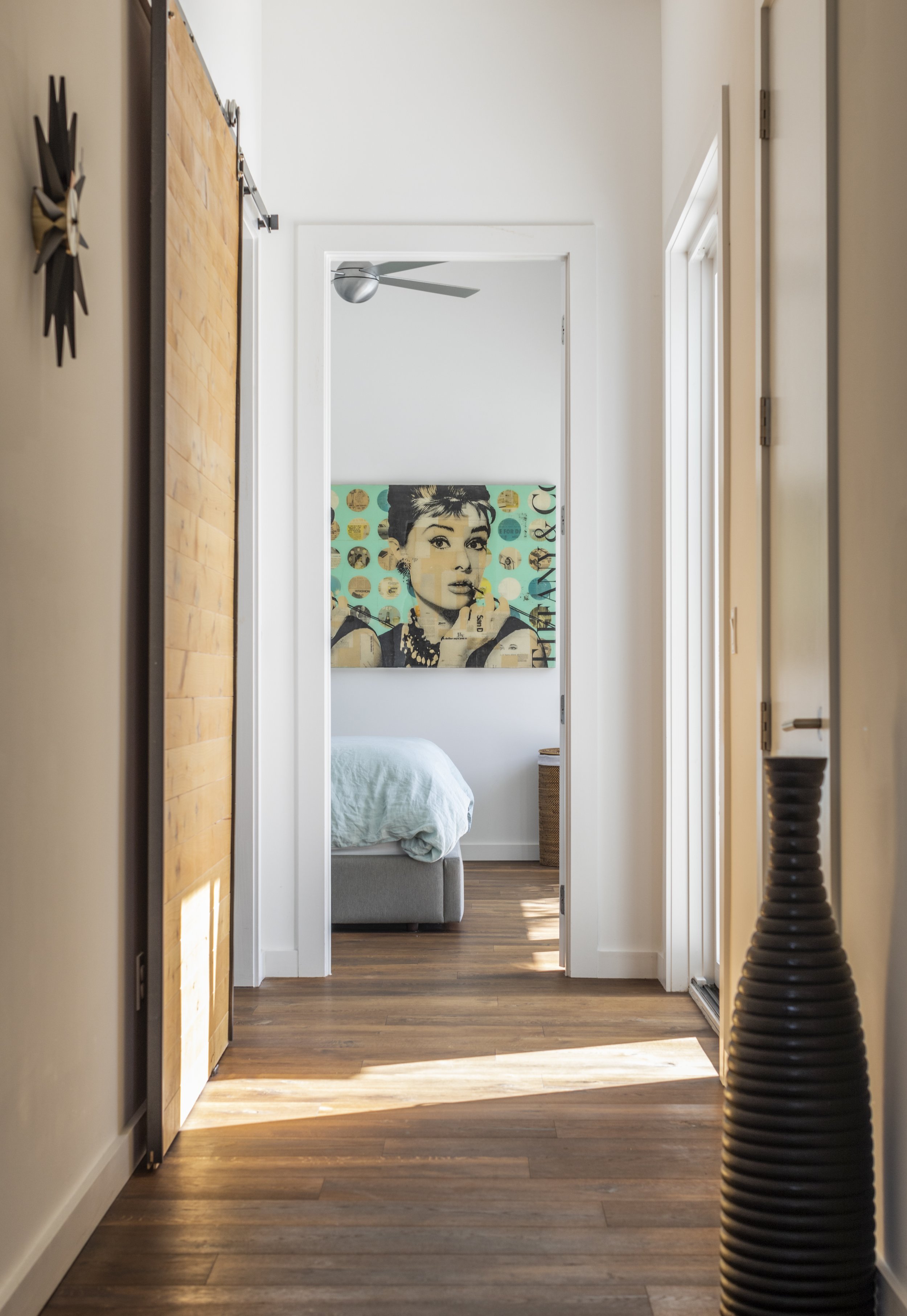 Empty hallway with wooden floor, white walls, and a view into a bedroom with a bed and a wall art of a woman.