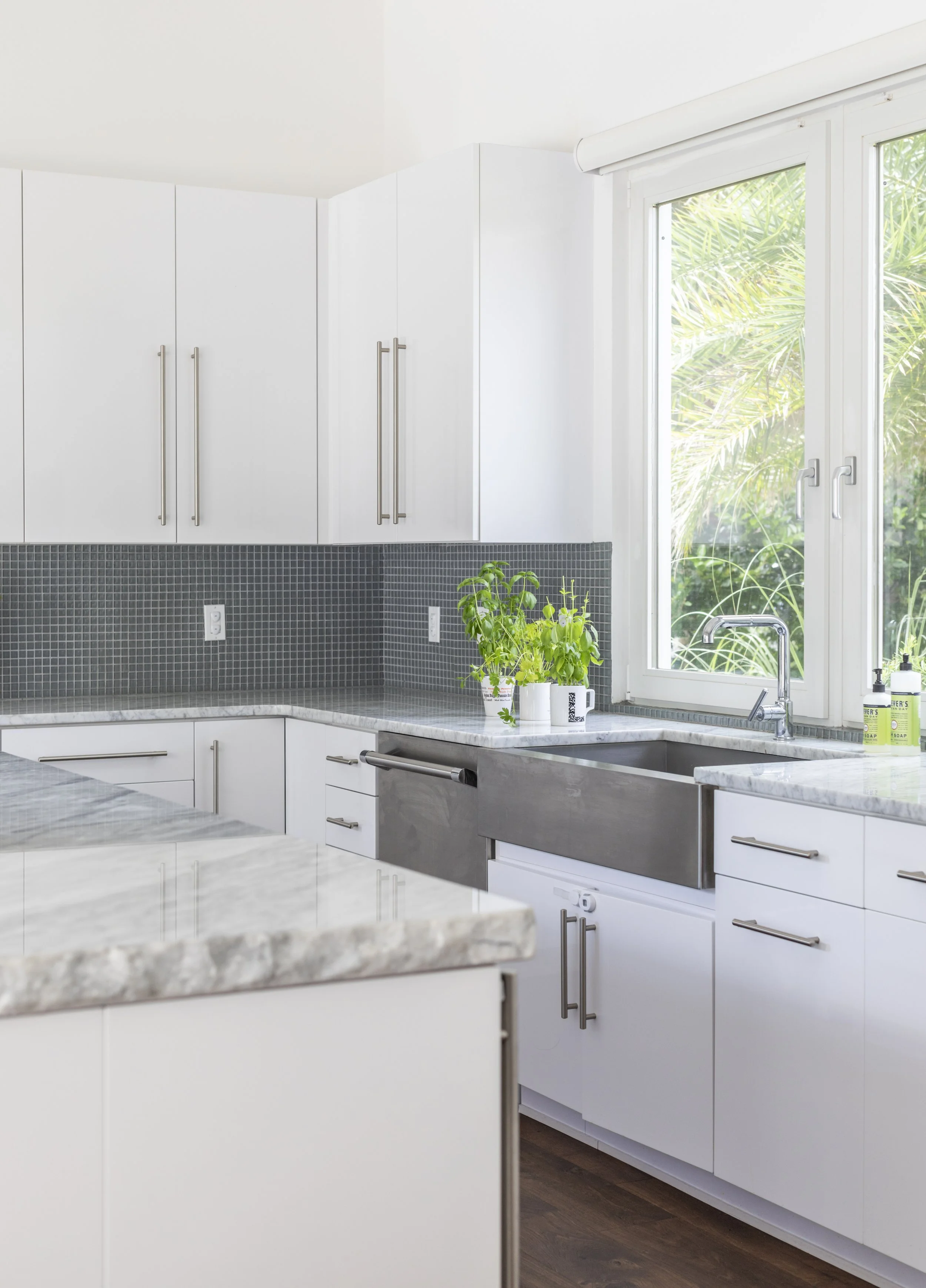 Modern kitchen with white cabinets, black tiled backsplash, marble countertops, a sink near a large window, and plants on the counter.