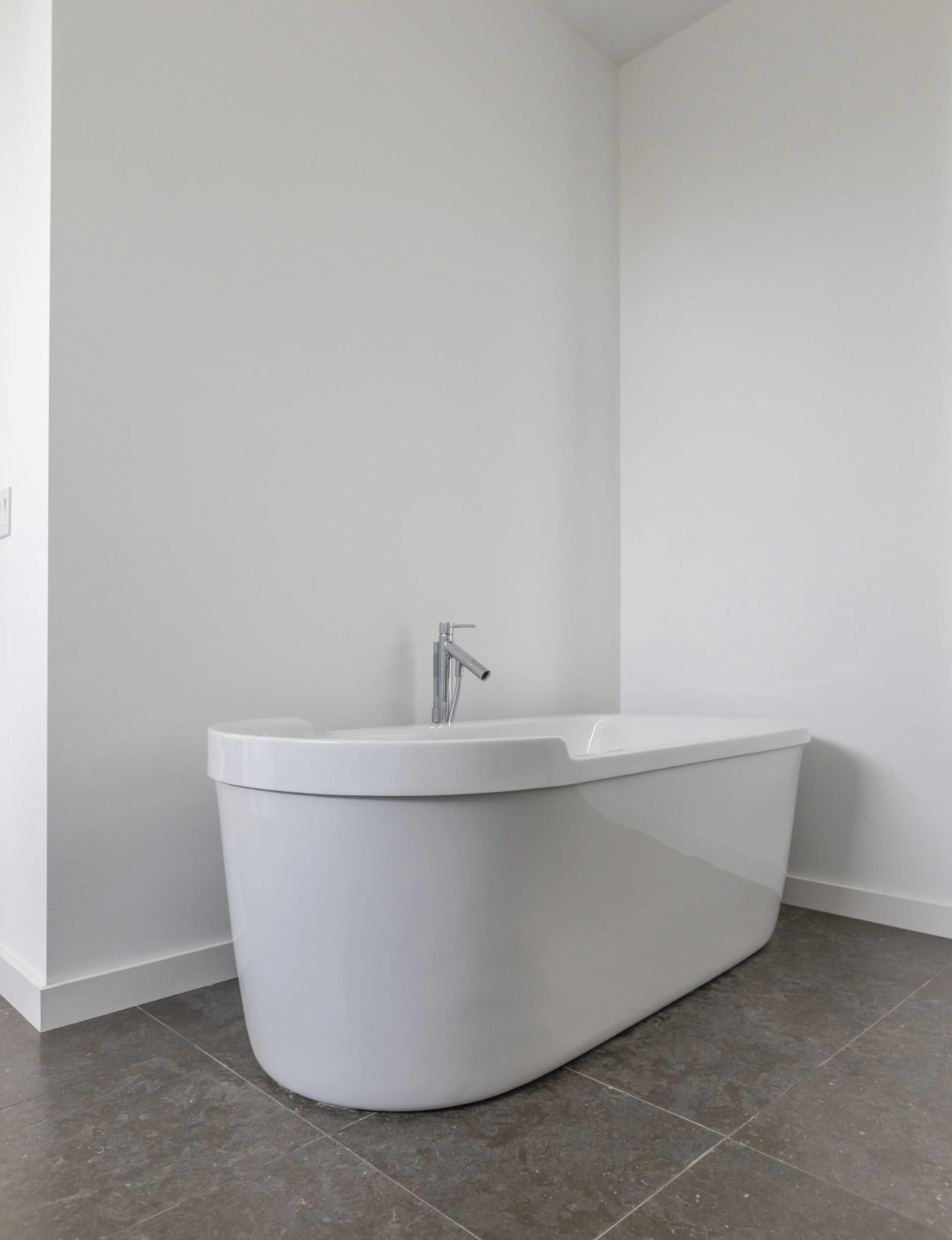 Modern white bathtub with a silver faucet in a bathroom, gray tiled floor, white painted walls, and a partial view of a laundry or utility area.