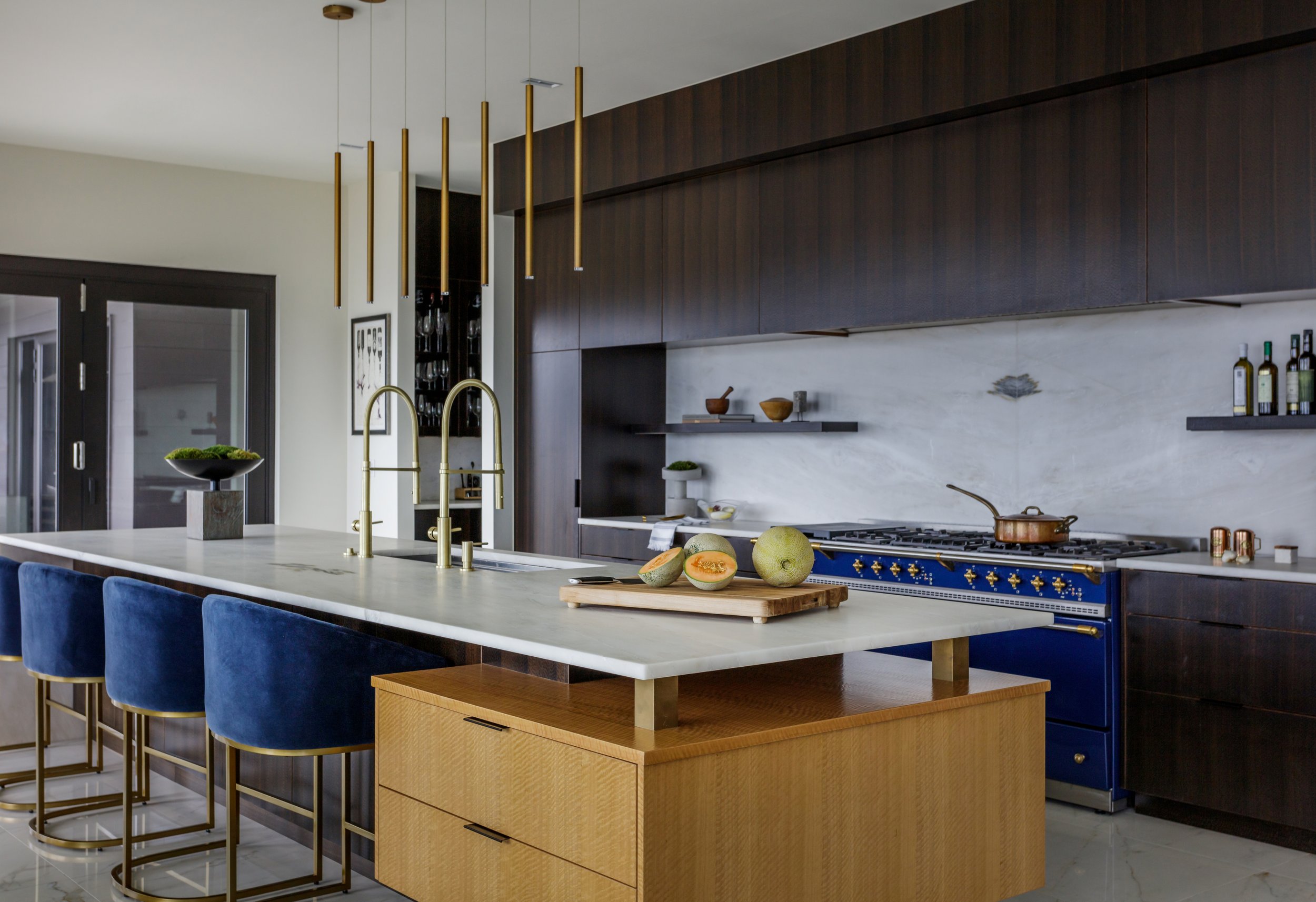 Modern kitchen with white marble island, blue velvet chairs, dark wood cabinets, gold pendant lights, and fresh fruit on the counter.
