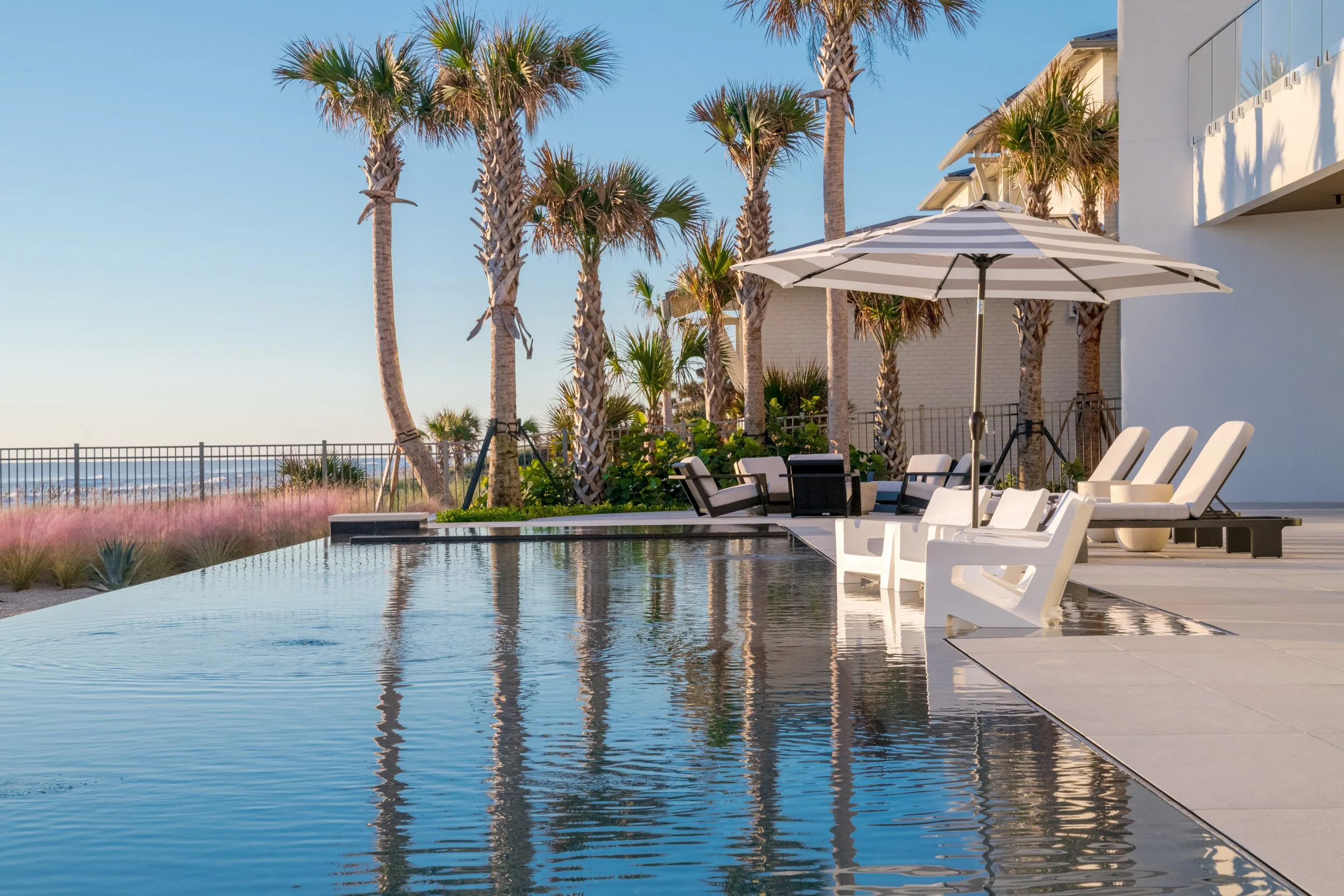 Poolside lounge chairs with a large striped umbrella, palm trees, and a modern building next to a body of water under a clear blue sky.