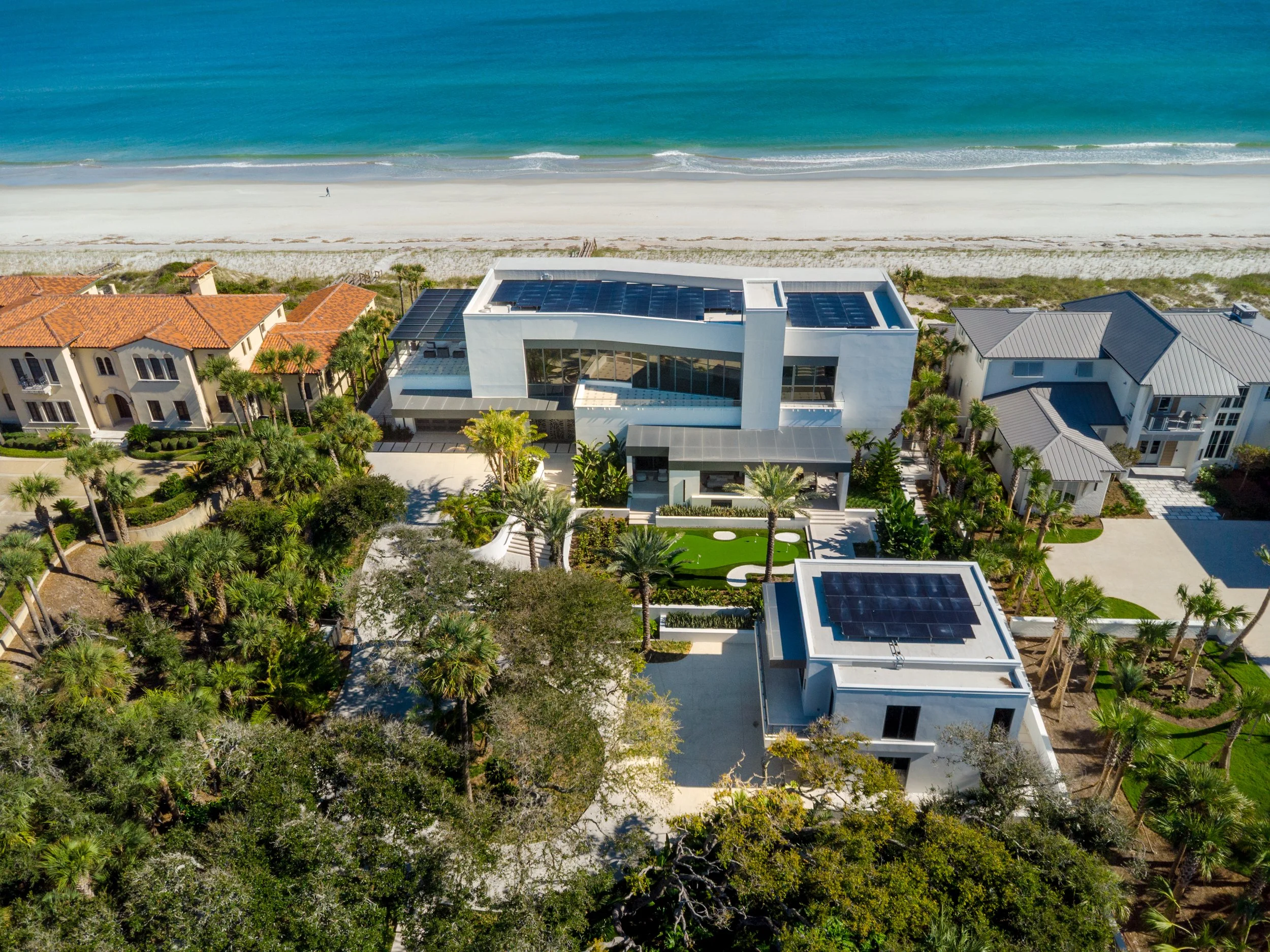 Aerial view of modern house with sunroof and solar panels, surrounded by palm trees, near a beach with white sand and turquoise water.