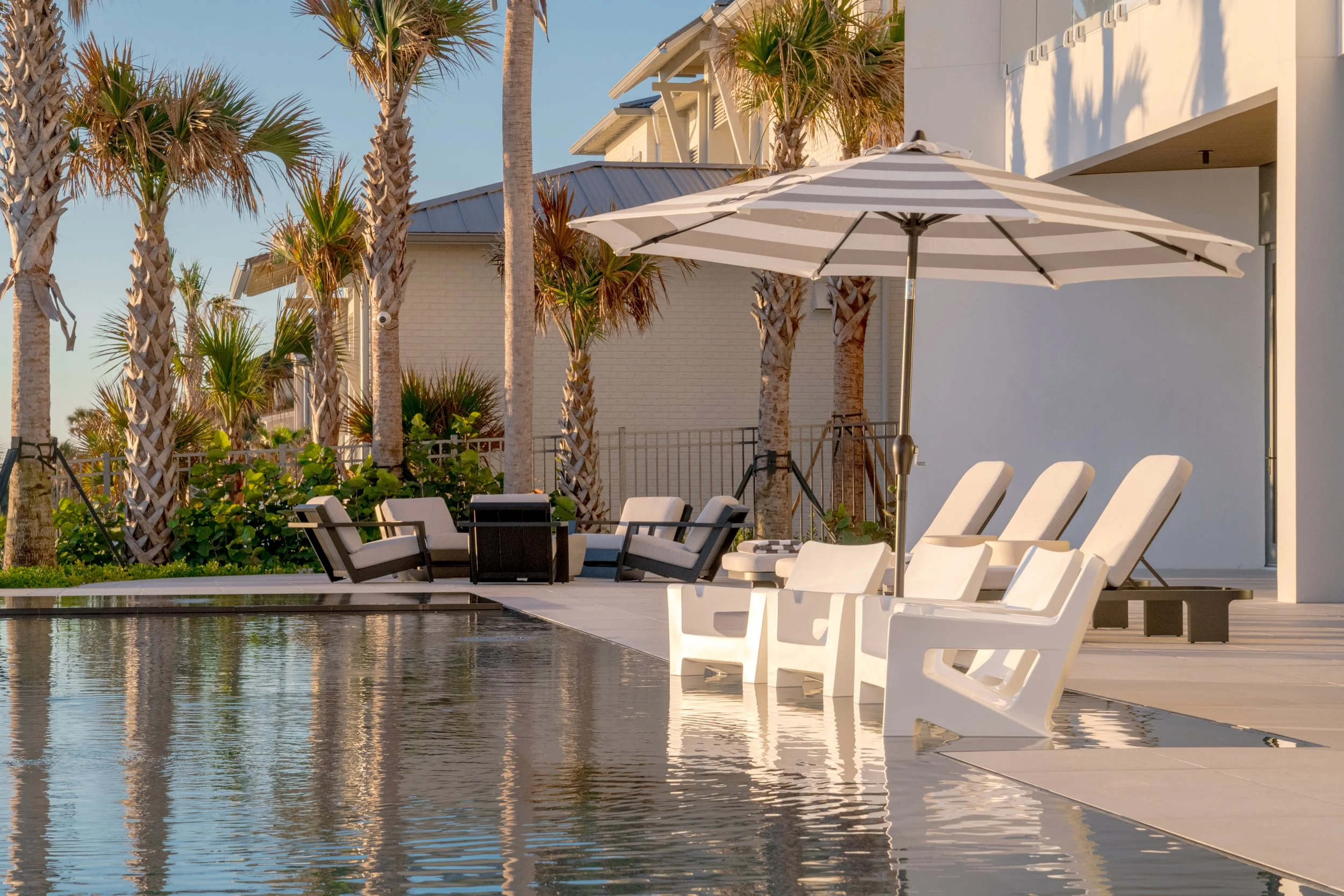 Poolside area with white lounge chairs, an umbrella, and palm trees near a modern house during daylight.
