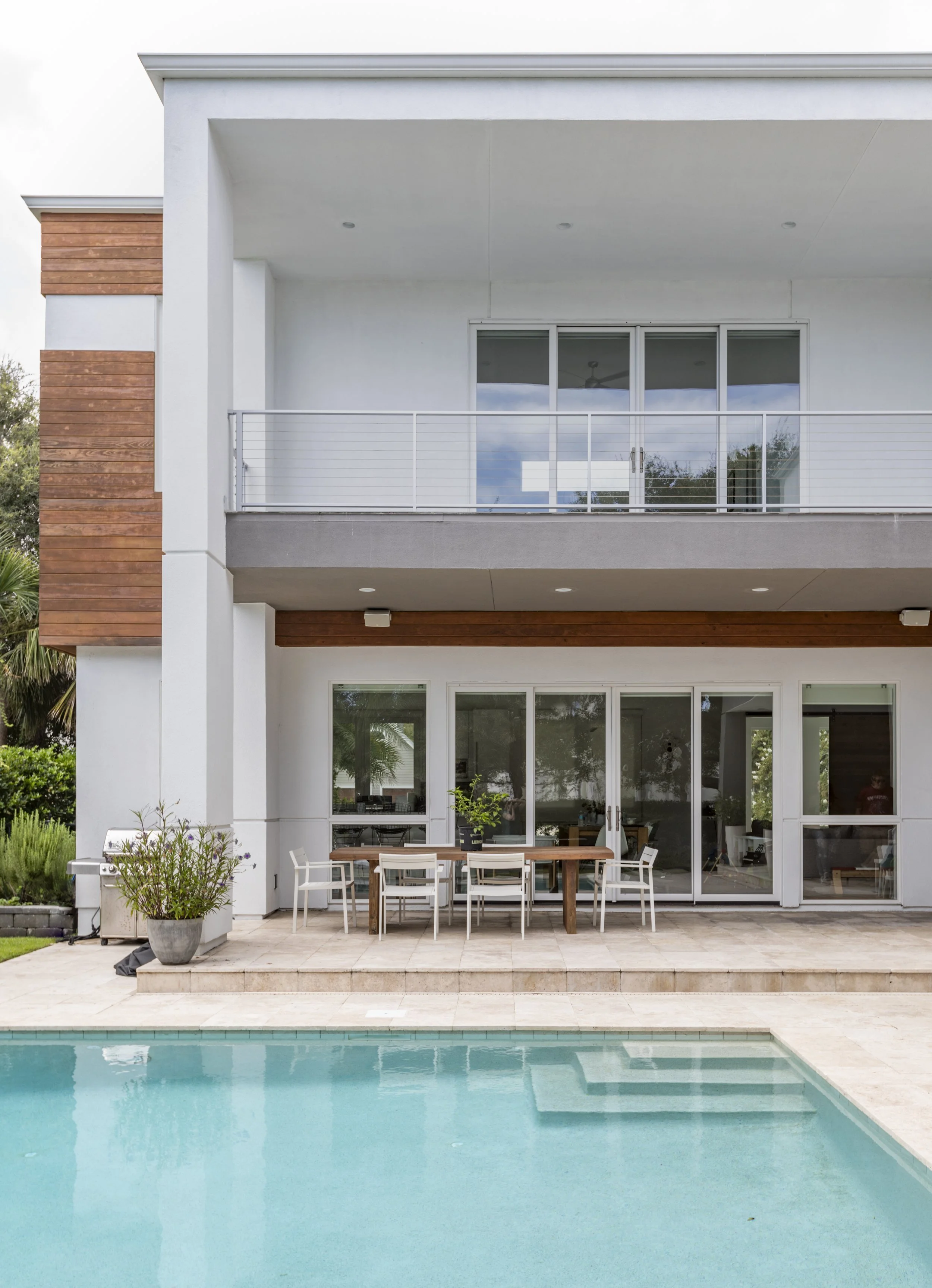 View of a modern house with a swimming pool. The house has two levels, large sliding glass doors, and a balcony with a white railing. There is a patio area with a wooden table and white chairs, and a potted plant on the patio. The pool is in the fore