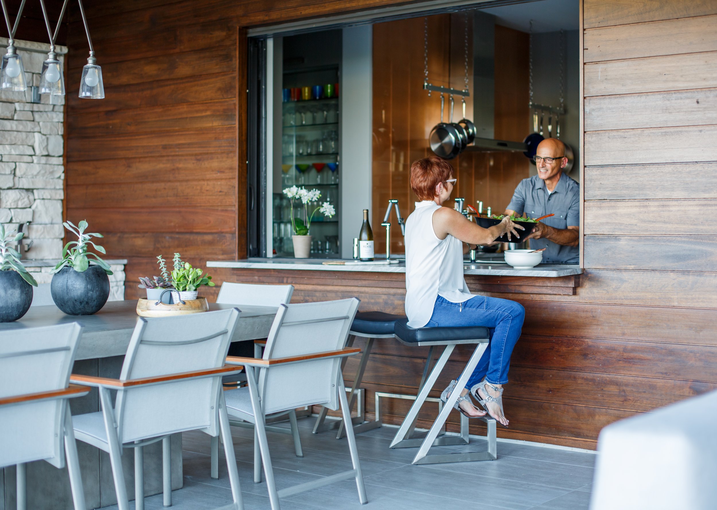 An older man is smiling and handing a salad to a woman with red hair. They are at a kitchen counter, with hanging pots and a wine bottle nearby. The scene is viewed from a dining area with white chairs and potted plants in the foreground.