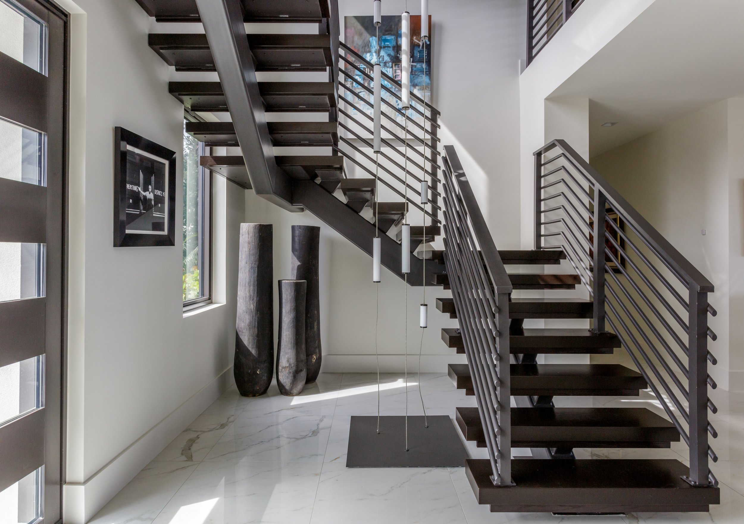 Modern interior staircase with dark wood steps, metal railing, hanging pendant lights, marble floor, and decorative tall vases by the window.
