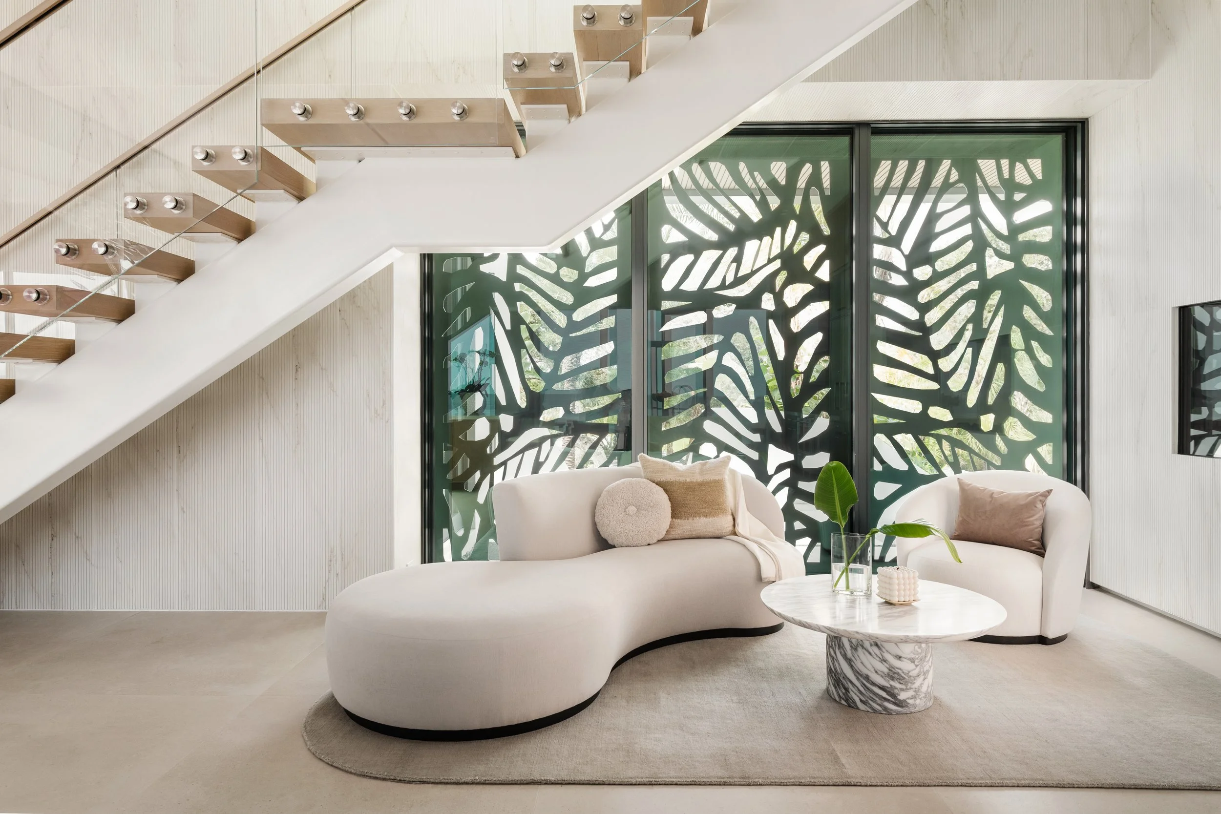 Modern living room with white curved sofa, marble coffee table, decorative pillows, large window with black grid frame and decorative green trees outside, staircase with wooden steps and glass railing.