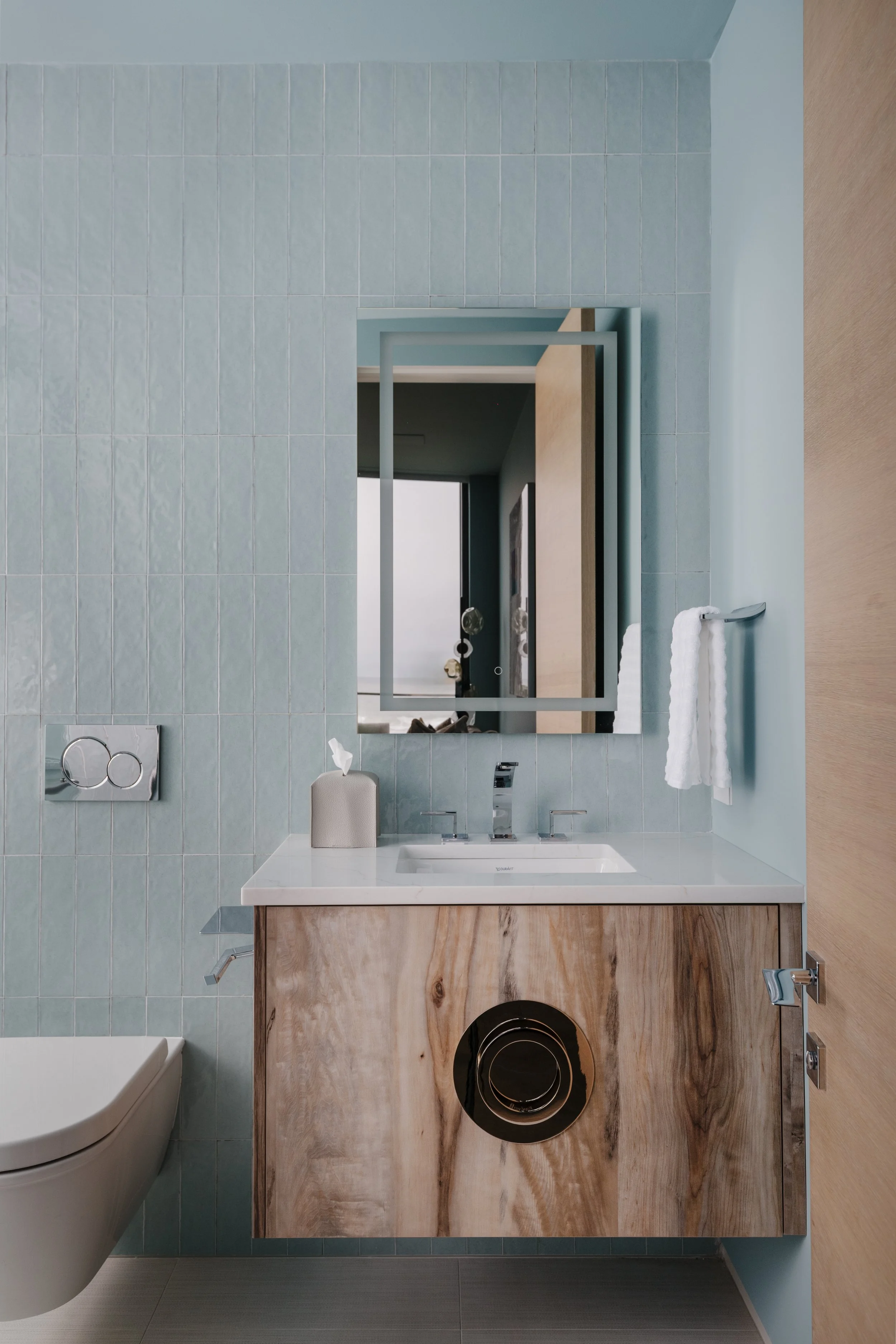 A bathroom interior with light blue tiled wall, a wooden vanity with a white countertop, an integrated sink, a large mirror, a white hand towel on a rack, and a washing machine with a black circular door beneath the counter.