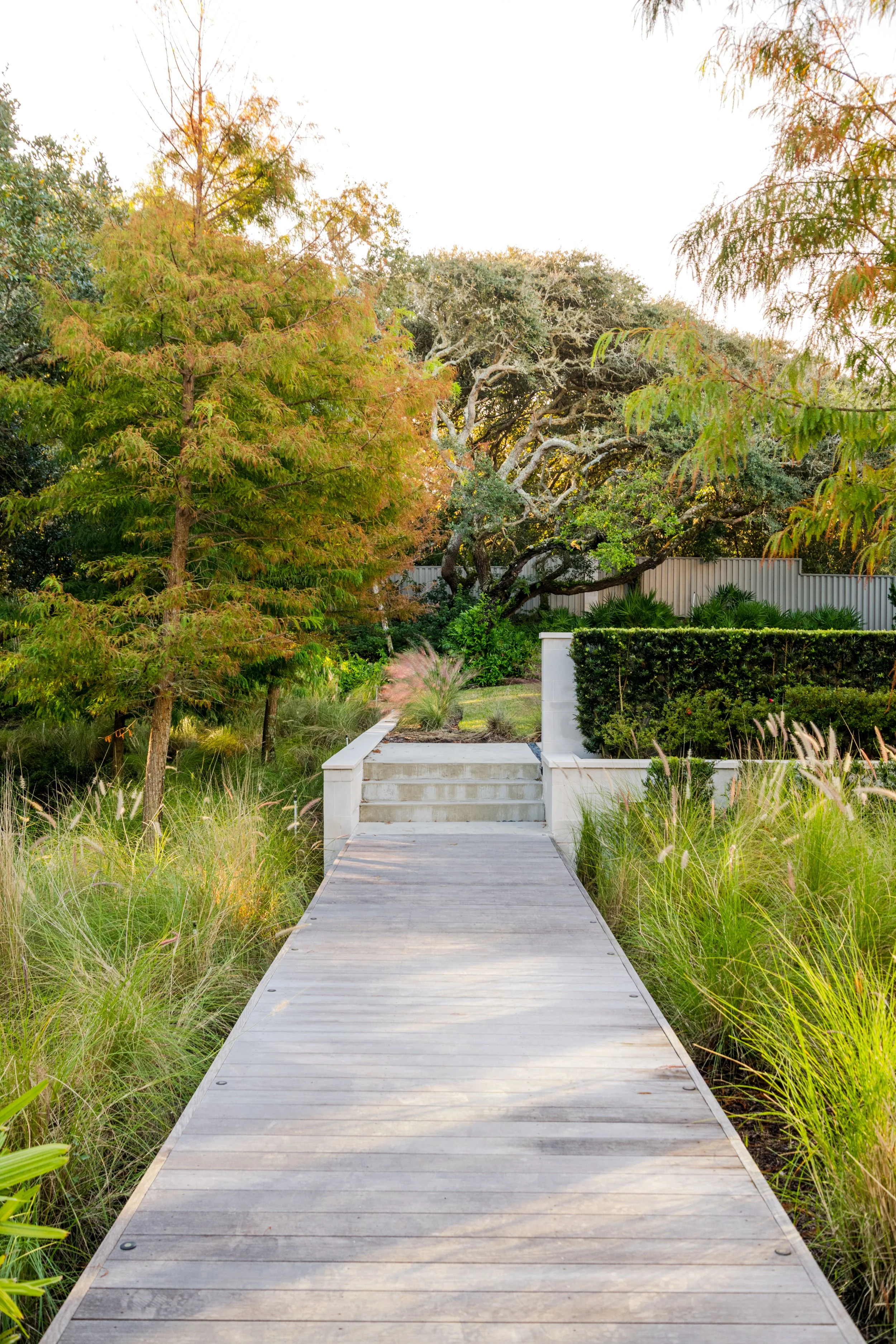 Wooden pathway leading to a garden with lush green plants, trees with autumn foliage, and a white fence in the background.