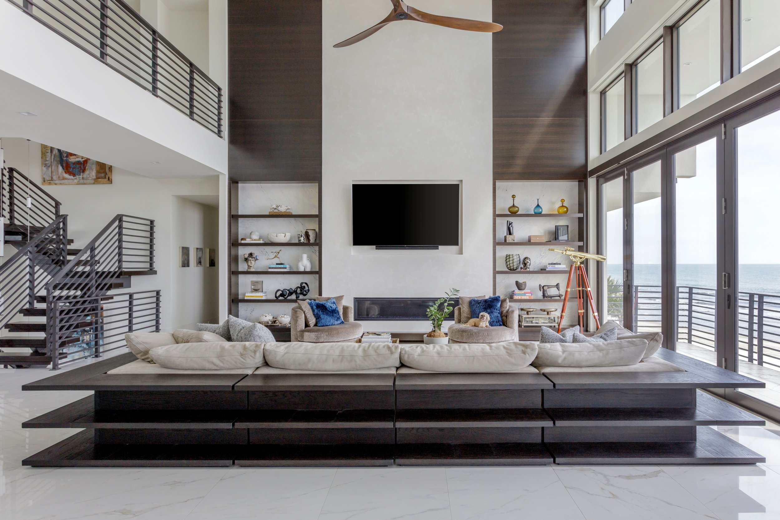 Modern living room with high ceilings, a large flat-screen TV on a white wall, built-in shelves with decorative items, a beige sectional sofa with blue pillows, a black coffee table, and large windows with ocean views.