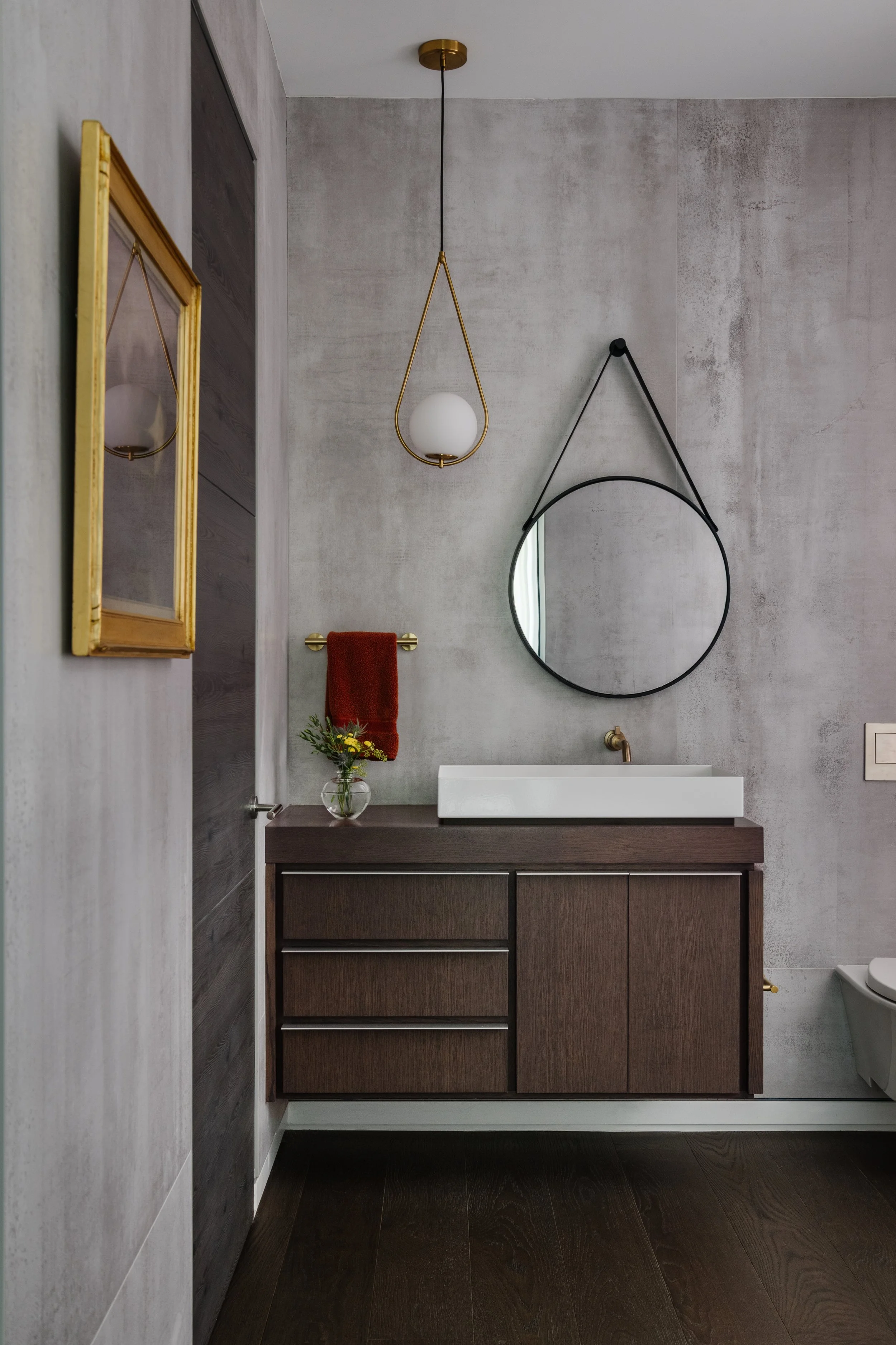 Modern bathroom with a dark wood vanity, white vessel sink, round mirror hanging with black strap, gold faucet, red towel, hanging white globe pendant light, textured gray wall, and dark wooden flooring.