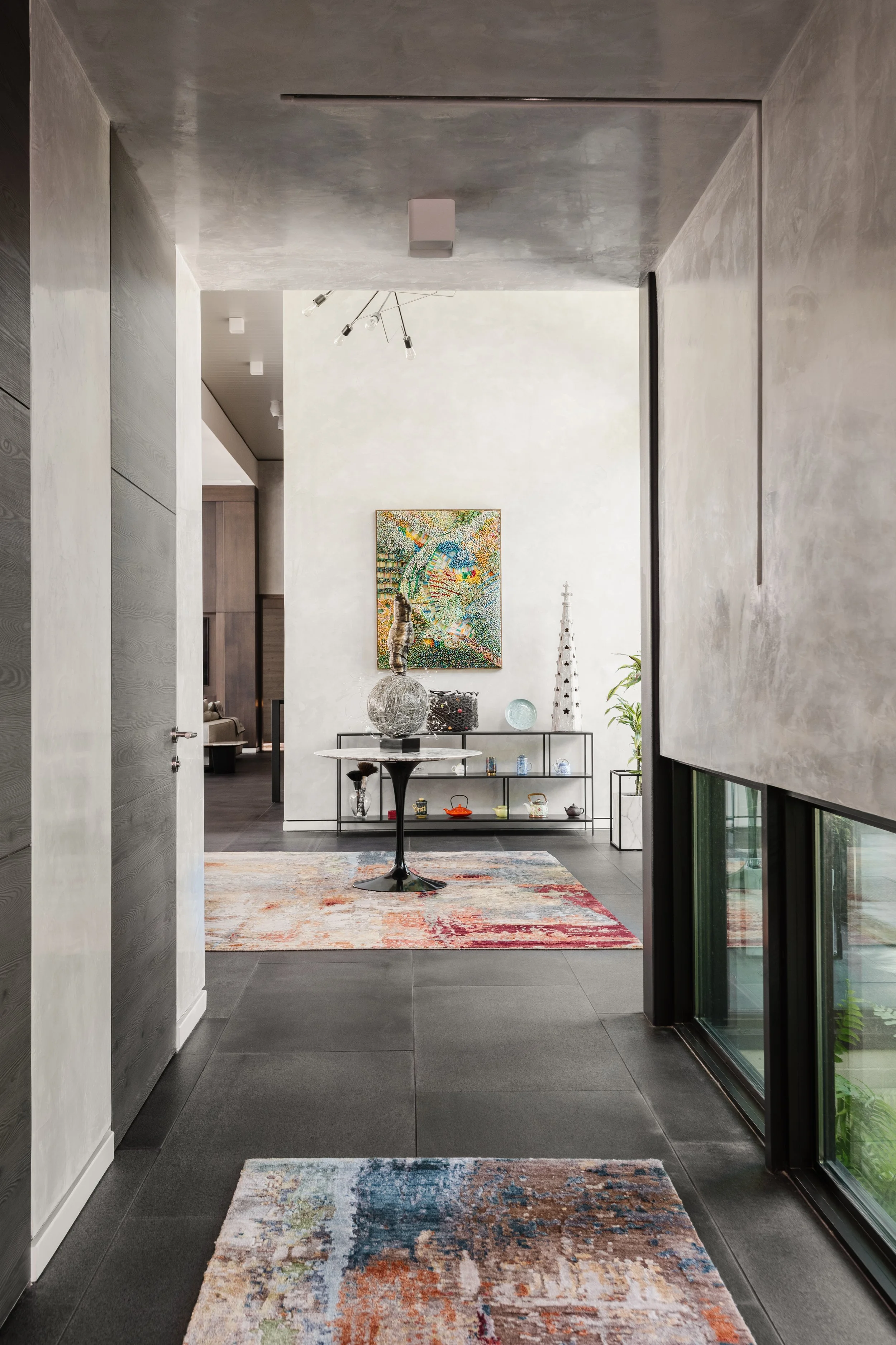 Modern interior hallway with a colorful abstract rug, a glass-topped table with decorative objects, and a white wall with a piece of colorful artwork. There is a black metal shelving unit with ceramics and glassware, and a tall white sculpture next t