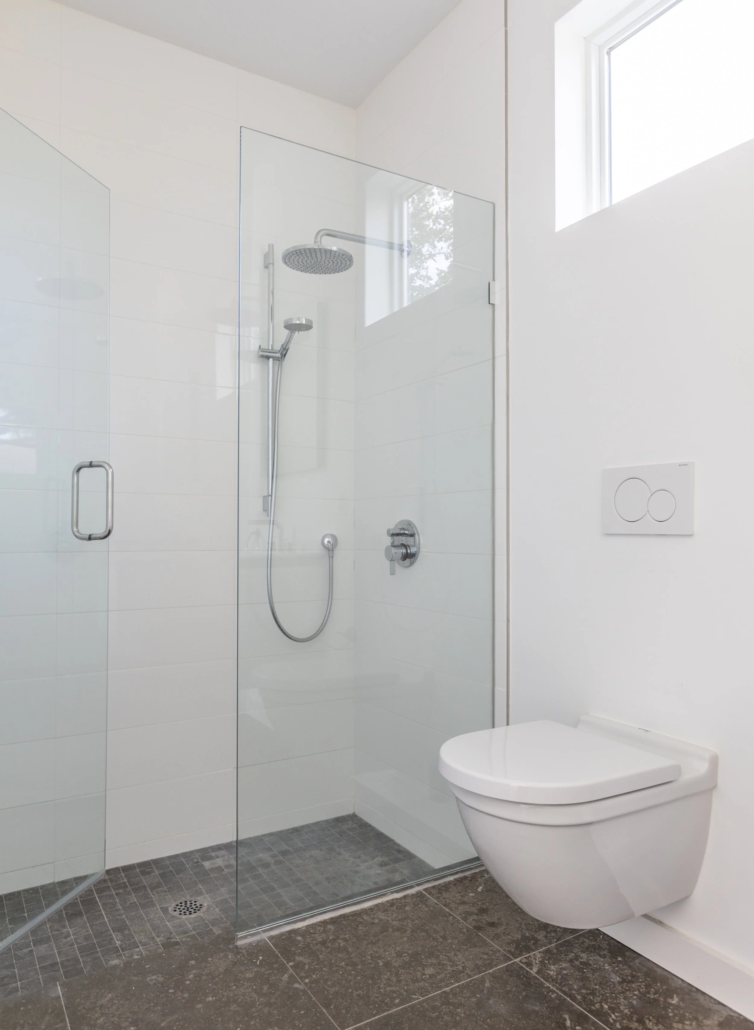 Modern bathroom with a glass shower enclosure, contemporary toilet, dark tile flooring, white walls, and a high window.