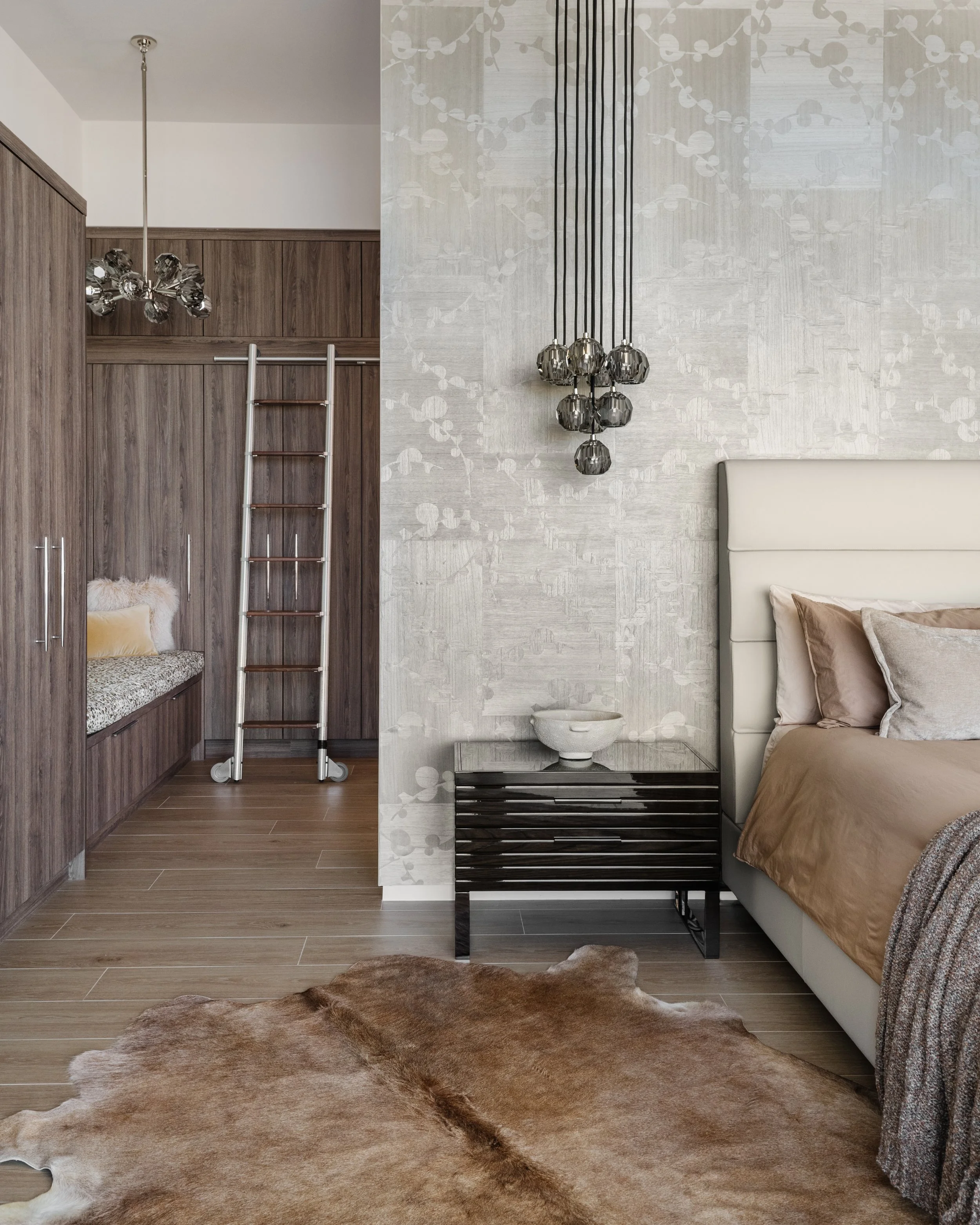 A bedroom with a portion of a bed, beige bedding, a black and white side table, a textured beige wall with floral patterns, a modern black chandelier, a white ceramic bowl, and a brown and white cowhide rug on a wooden floor. There is also a wall wit
