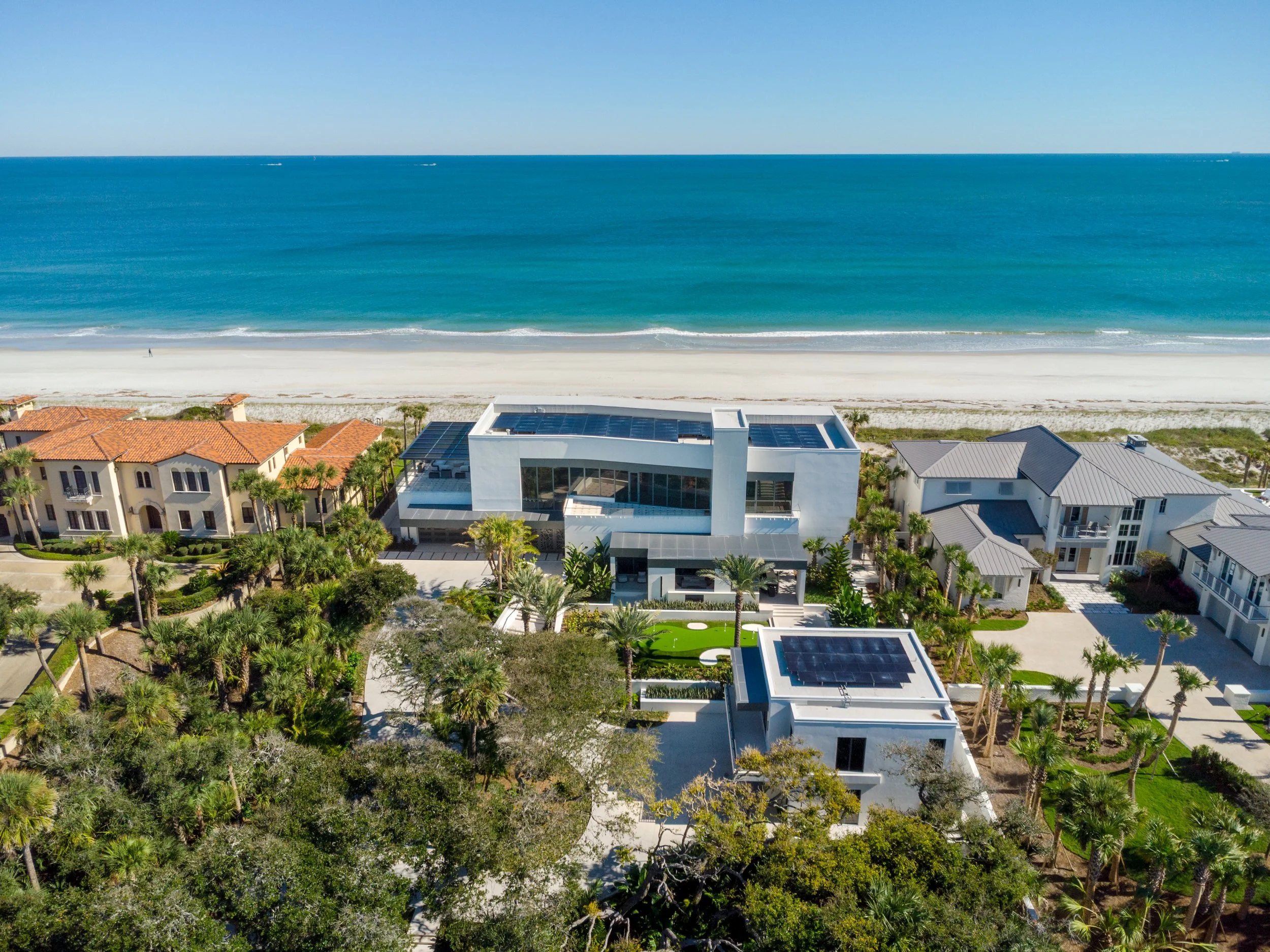 An aerial view of beachfront houses with red, gray, and white roofs, lush greenery, and a white sandy beach leading to turquoise ocean waters under a clear blue sky.