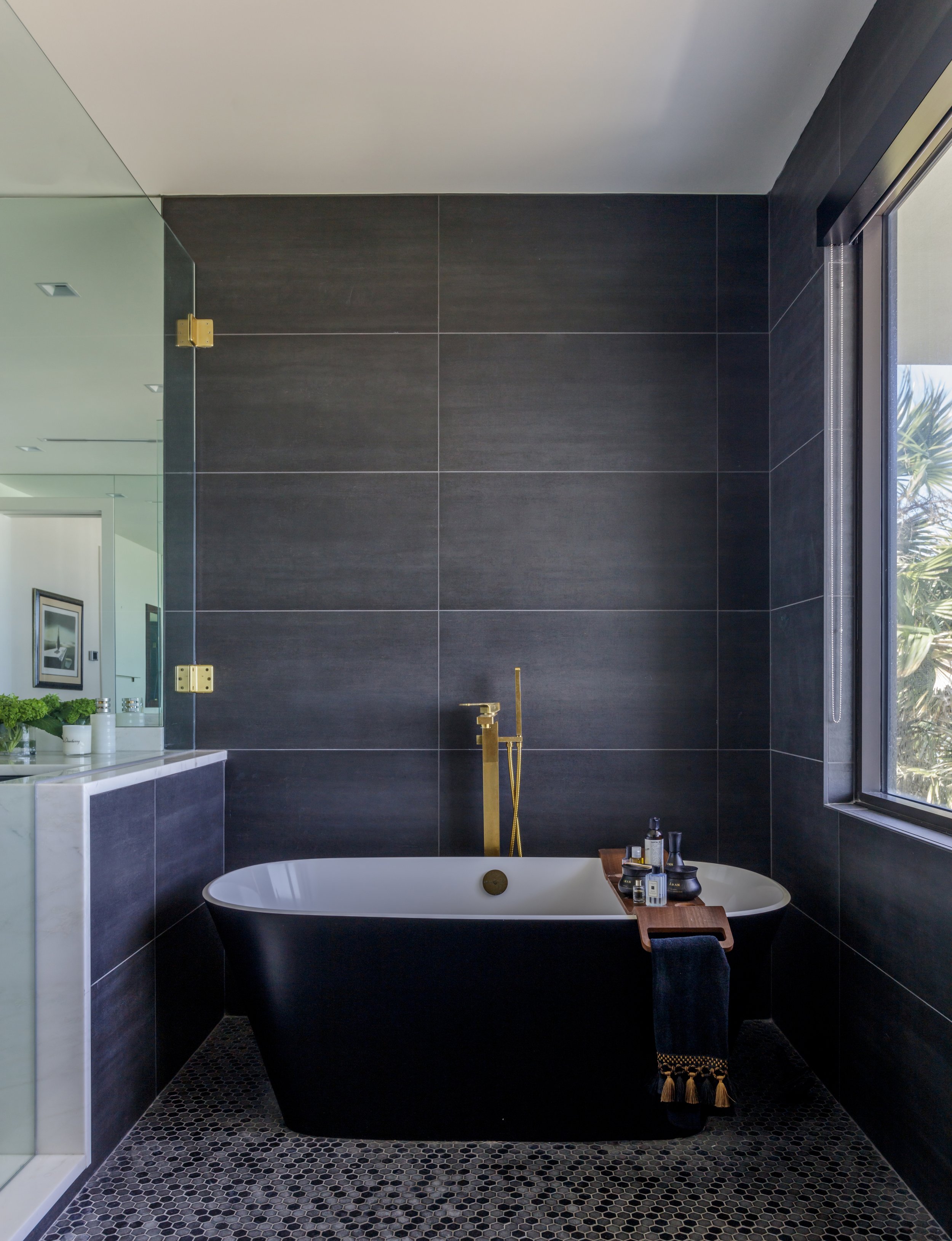 Modern bathroom with black tiled walls, a freestanding bathtub with a wooden tray, bottles of toiletries, a gold faucet, and a large window with a view of greenery outside.