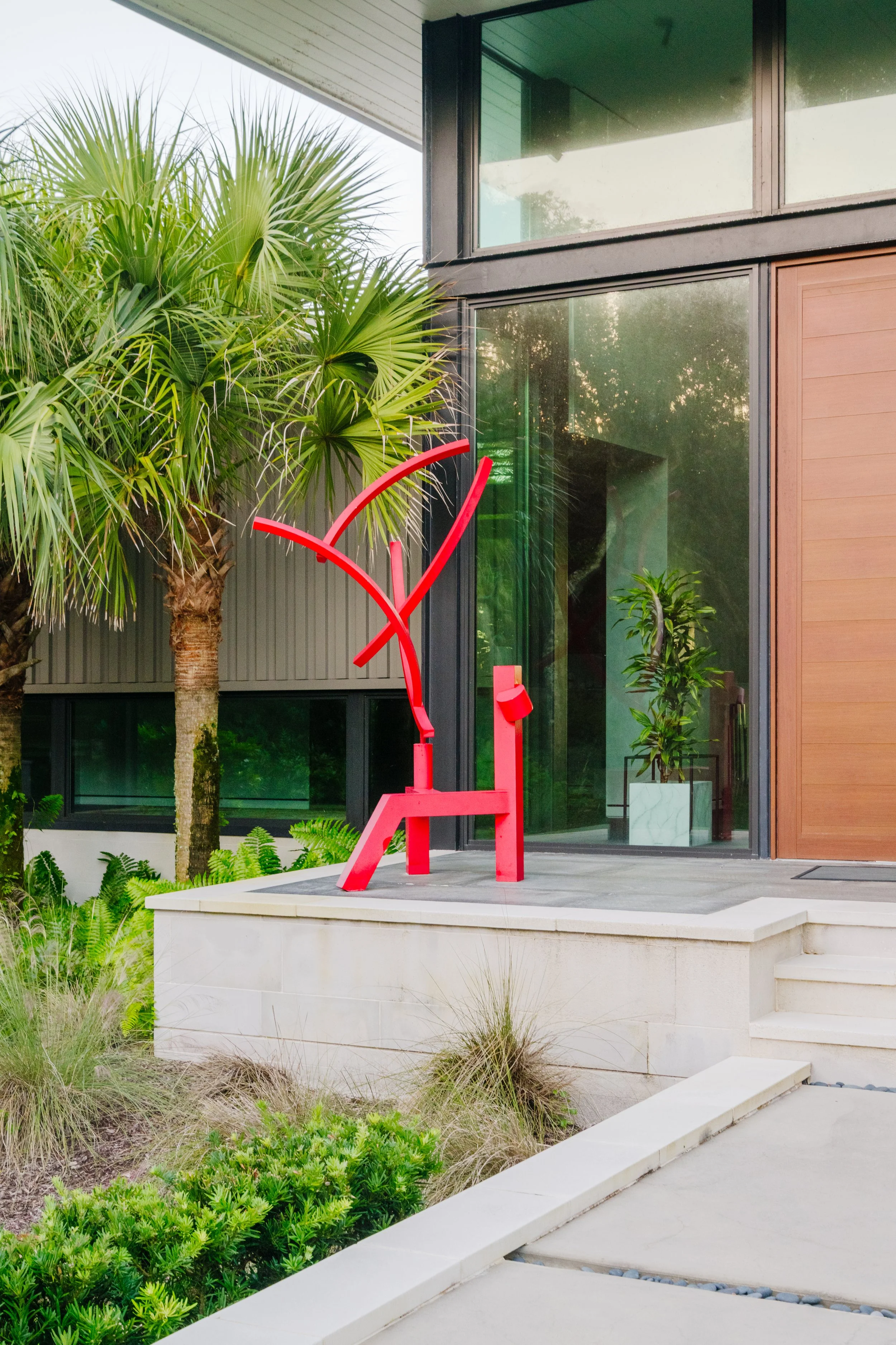 Red sculpture of a chair outside a modern house with large glass windows, surrounded by green plants and trees.