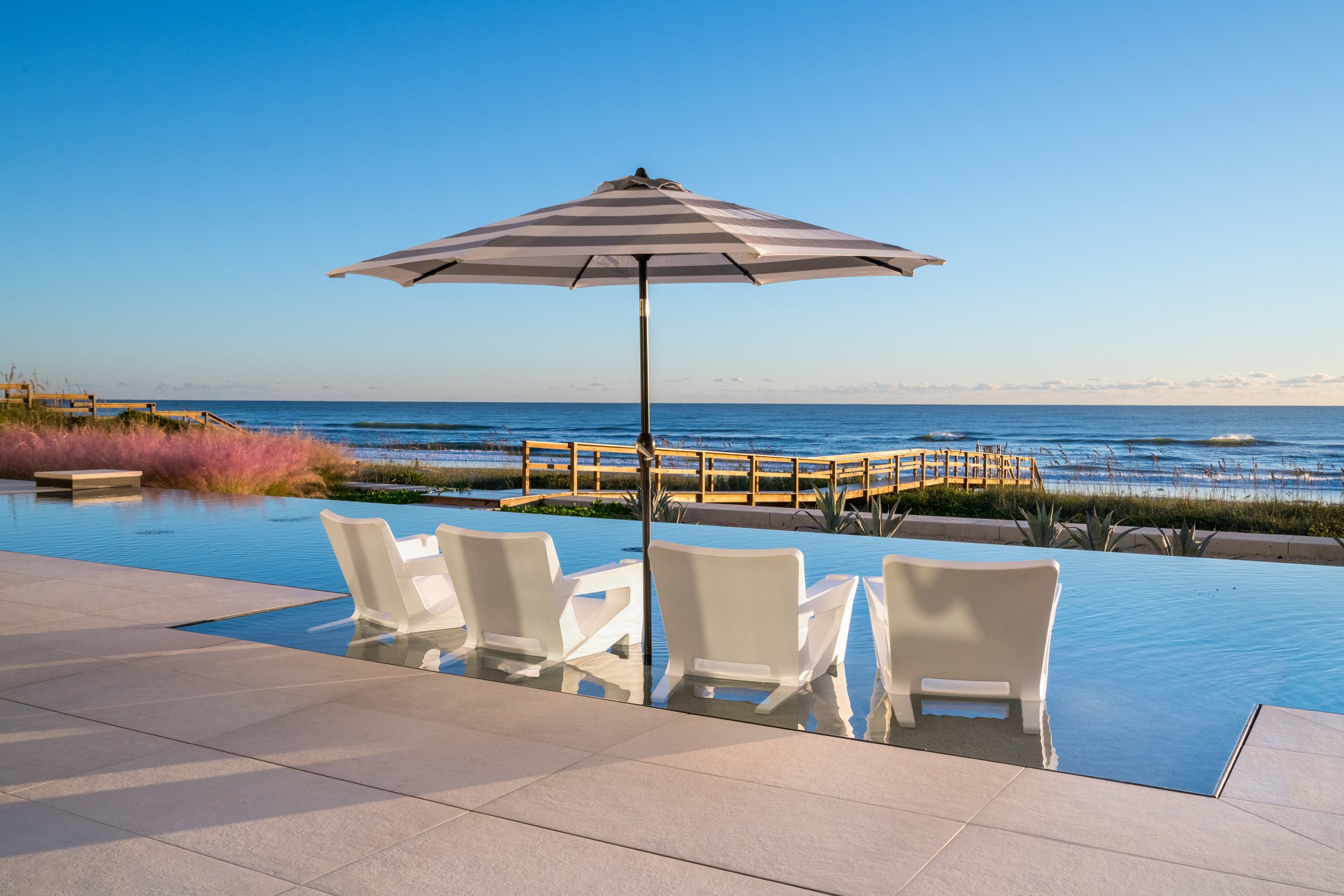 Outdoor poolside area with white chairs, a striped umbrella, and a view of the ocean in the background under a clear blue sky.