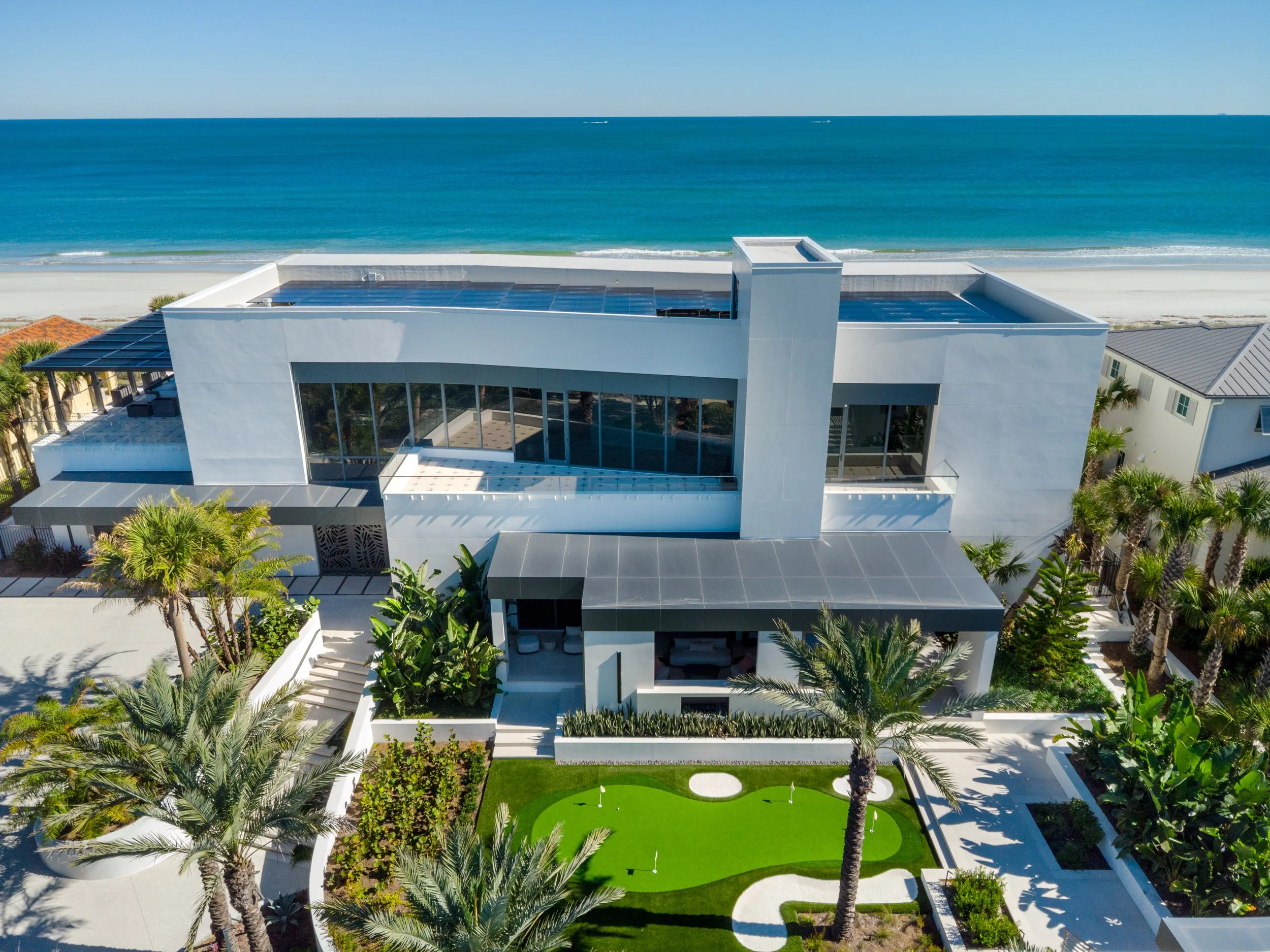 A modern, white, multi-story house with large glass windows and a rooftop pool, located by the beach with ocean in the background and lush tropical trees surrounding the property.