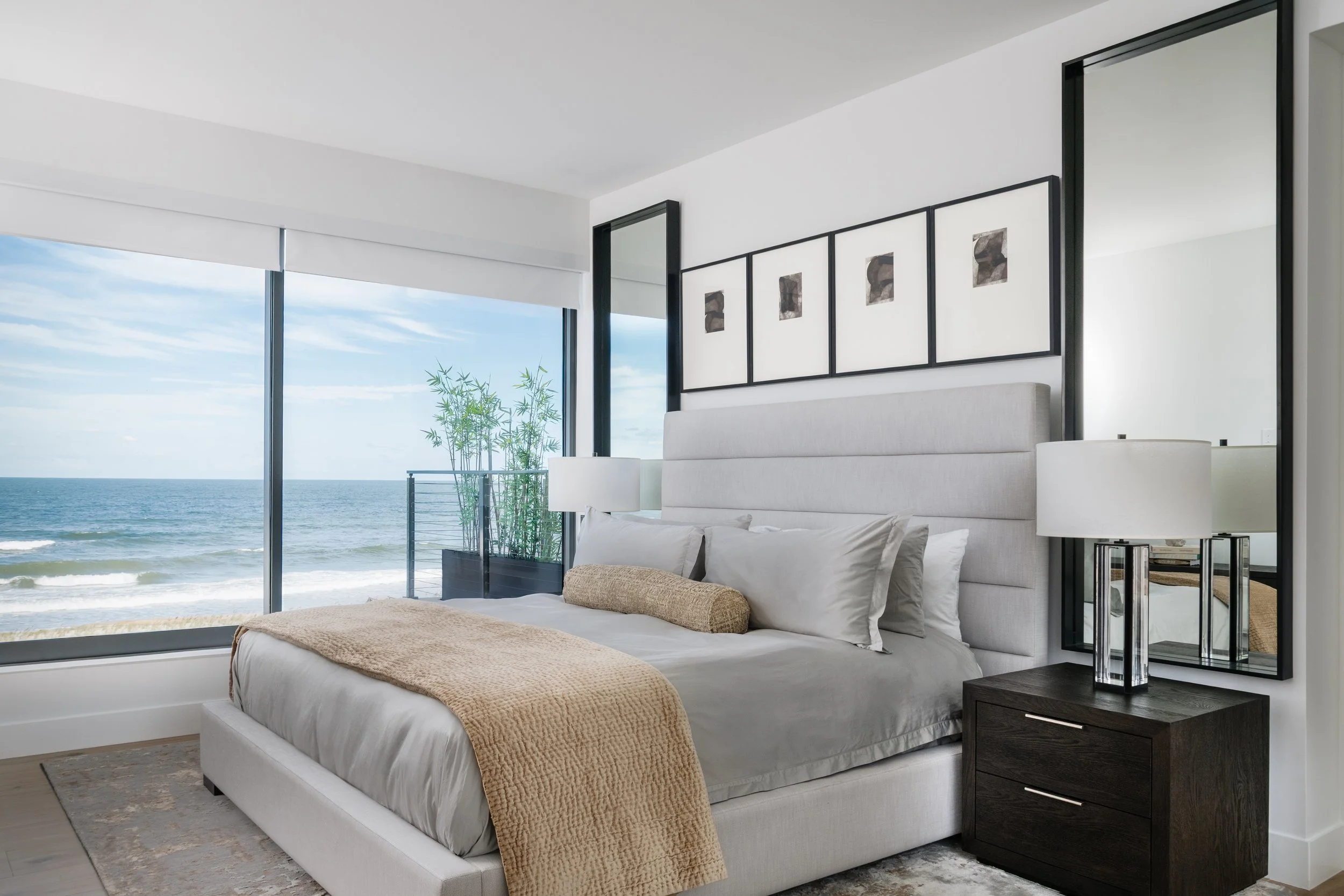 Modern bedroom with large windows showing ocean view, white bedding with beige throw blanket, black framed artwork on wall, nightstand with lamp and books, and a door leading to a balcony with plants.