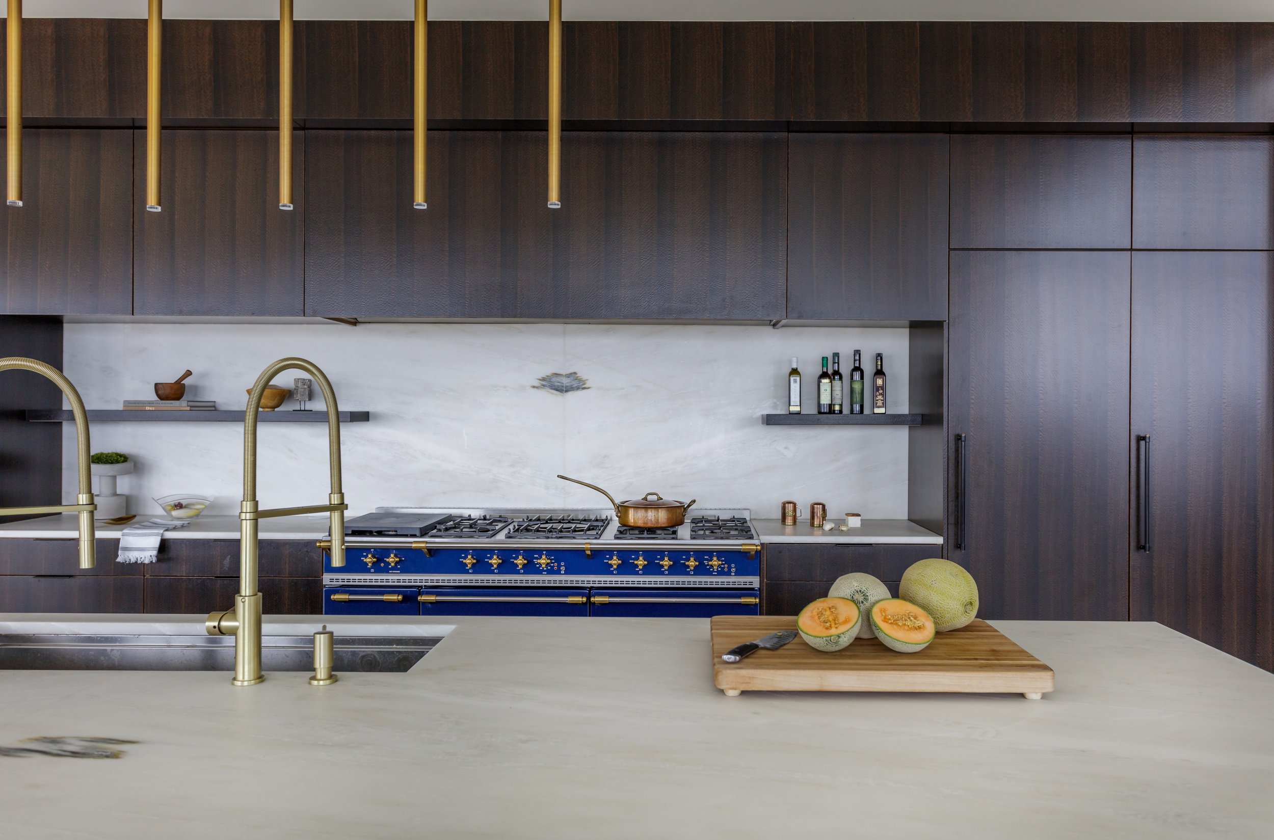 Modern kitchen with dark wooden cabinets, a white marble backsplash, and a blue and gold stove. A chopping board with sliced cantaloupe and a knife is on the white island counter near a gold faucet.