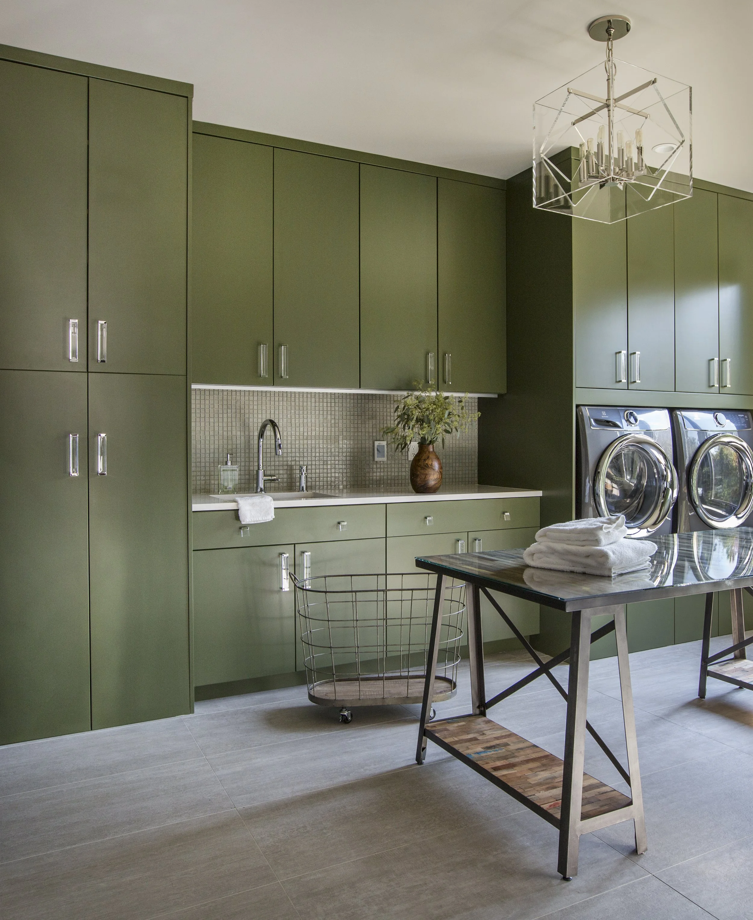 Green kitchen cabinets above a white countertop with a small sink, wood planter with flowers, and a black metal folding table with a stack of white towels and a wire basket chair in front of laundry machines.