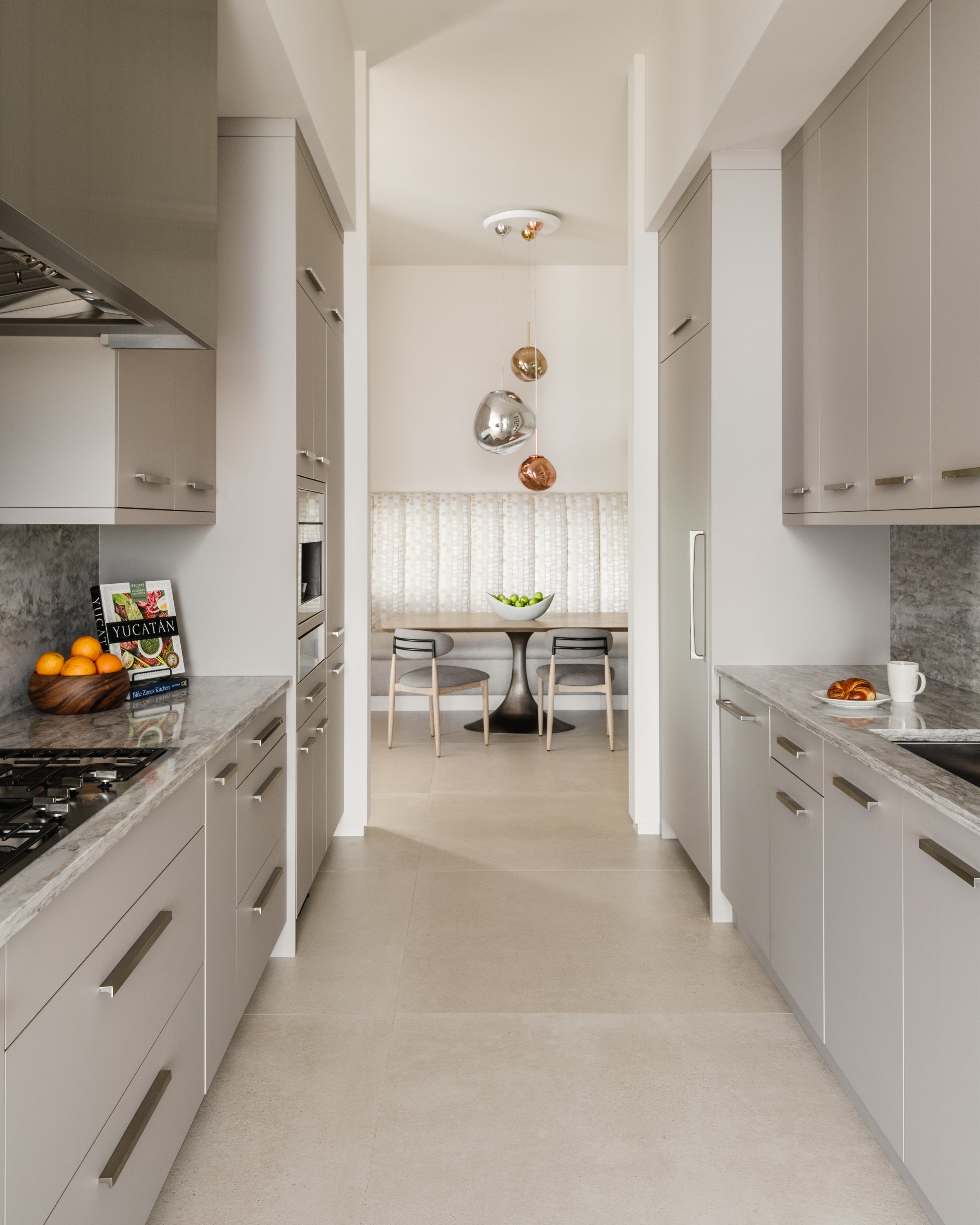 A modern kitchen with white cabinetry and marble countertops, view through a central corridor to a dining area with a round table, two chairs, a bowl of green apples, and a pendant light fixture hanging from the ceiling.