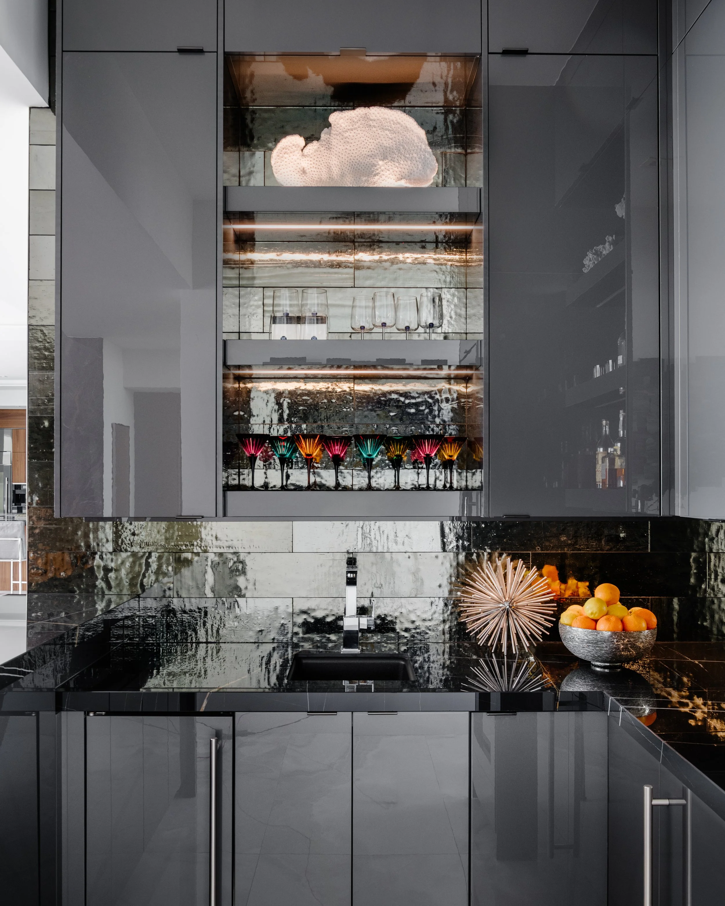 Modern kitchen with black cabinetry, marble backsplash, and decorative items including colorful glassware and a bowl of oranges.