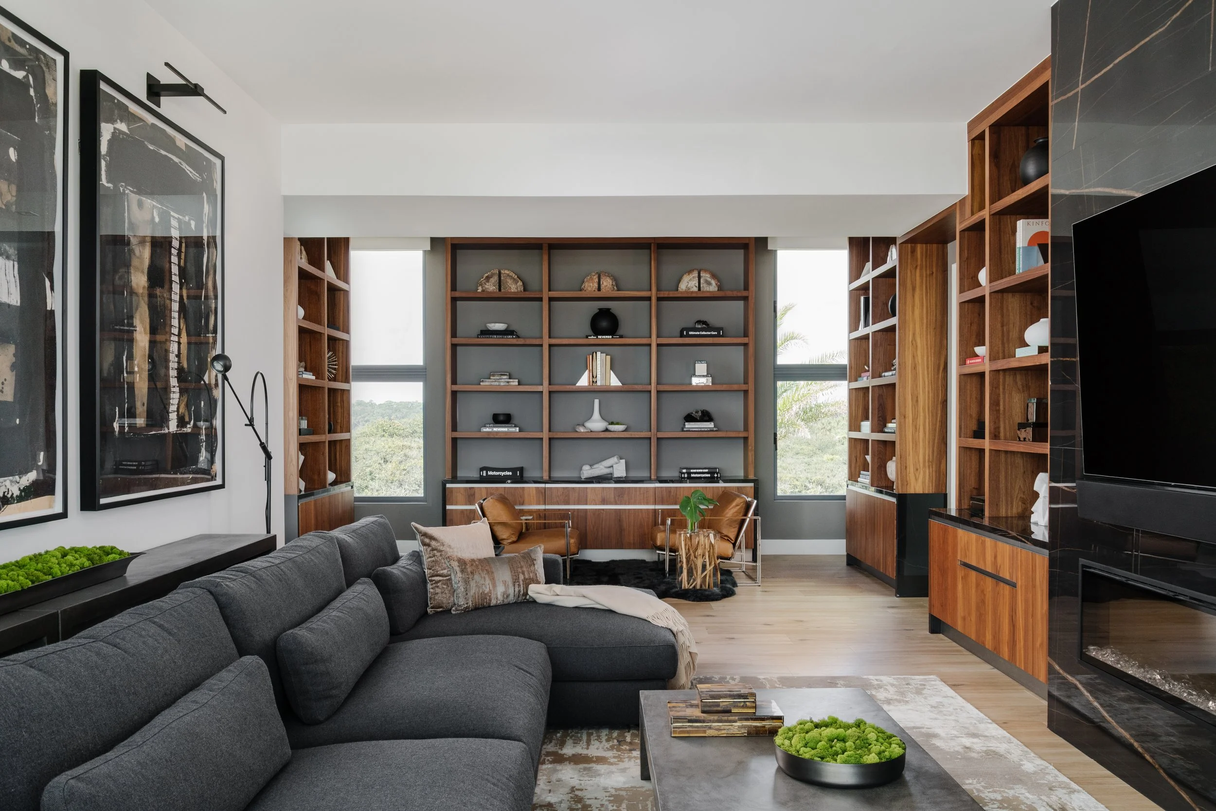 Modern living room with dark gray sofa, wooden bookshelves, abstract art, and large windows showing outdoor greenery.
