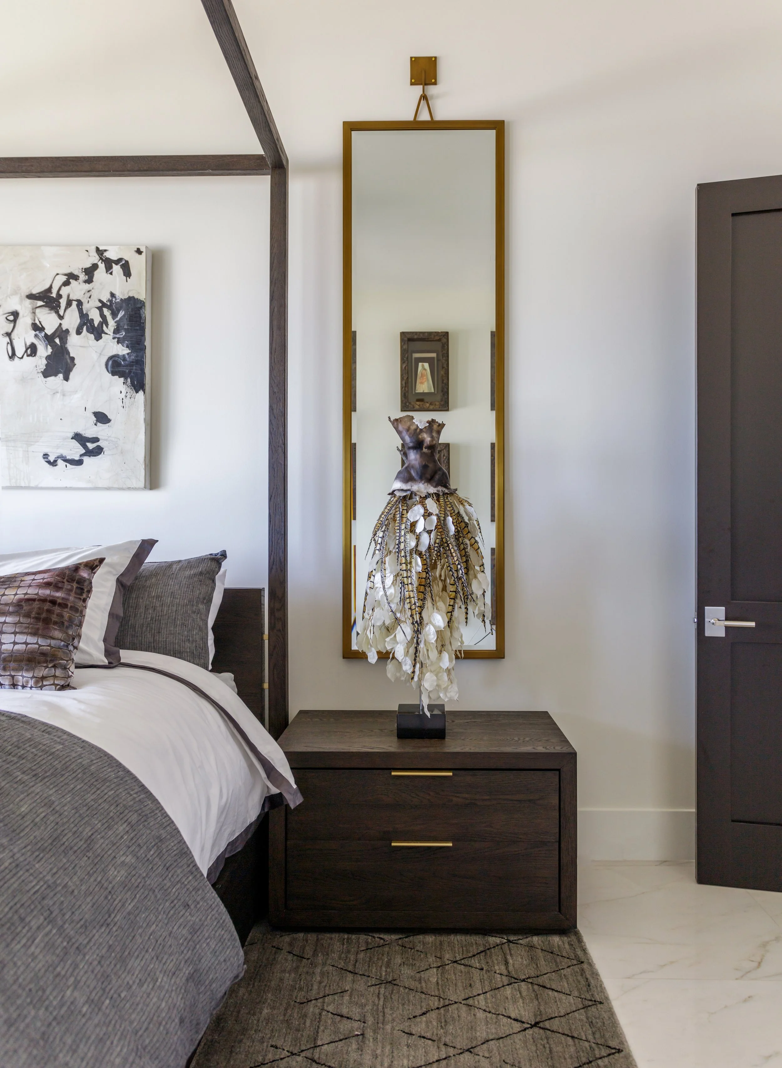 A bedroom corner with a bed, pillows, blanket, a dark wooden nightstand, a large mirror, a wooden sculpture, and framed artwork on the wall.