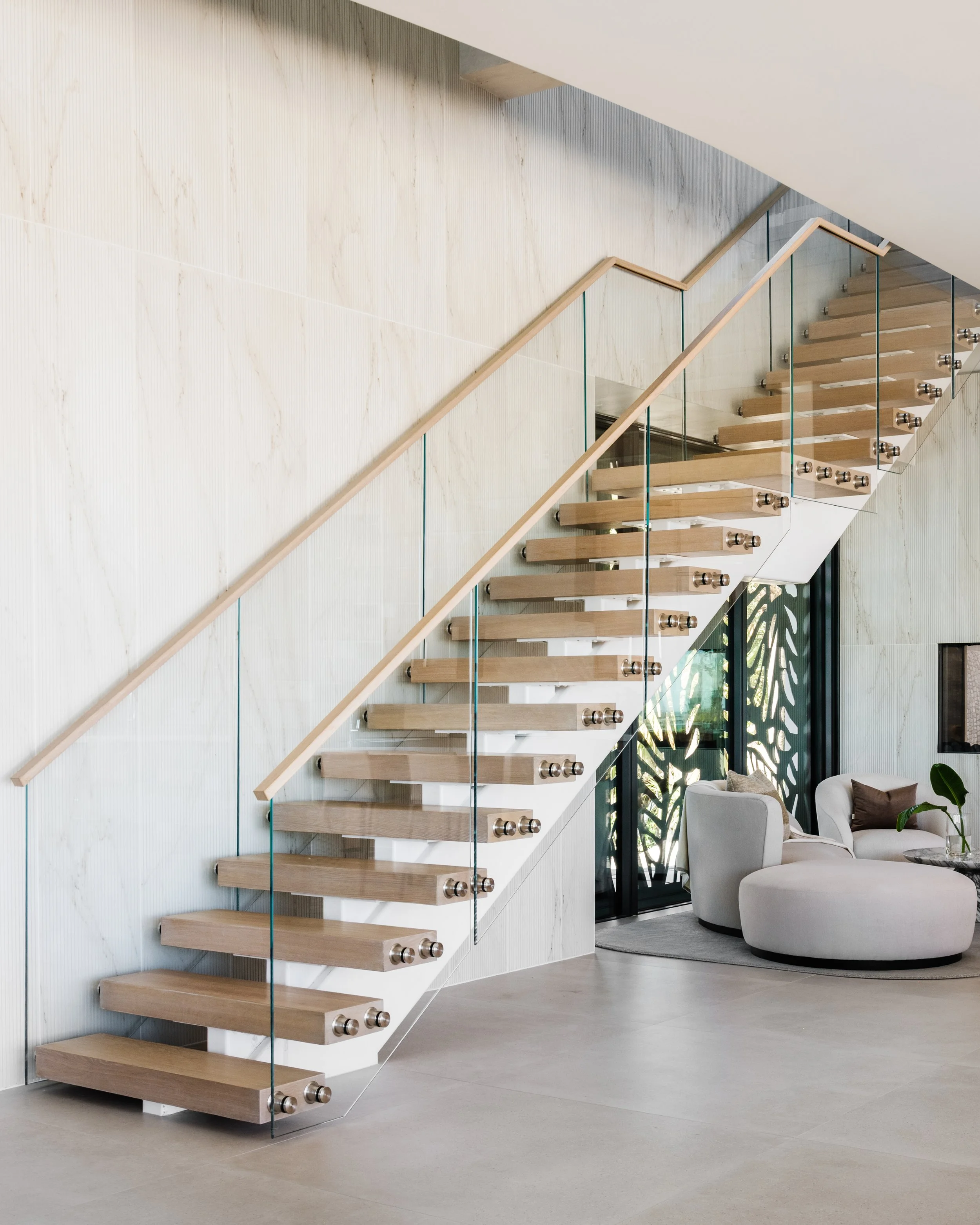 Interior of a modern house with floating wooden stairs, glass railings, a light-colored wall, and a view into a living room with a white sofa and glass doors leading outside.
